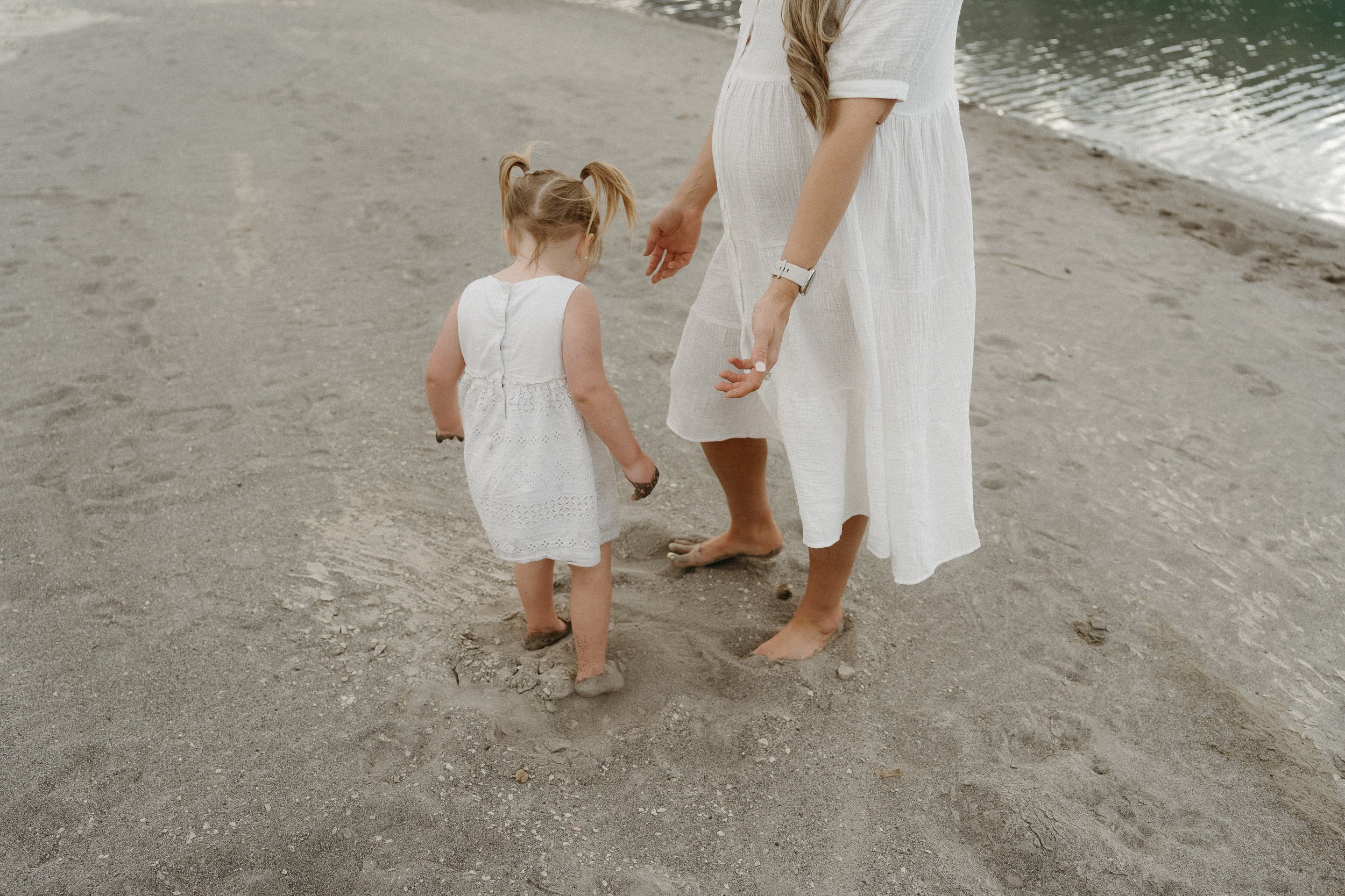 Mountain Maternity Session.  Calgary, AB.  mom and girl both dressed in white, walking barefoot on a sandy beach near the water, with the girl looking down at the sand. The scene has a peaceful and natural atmosphere. The text 'everyday life". Banff 