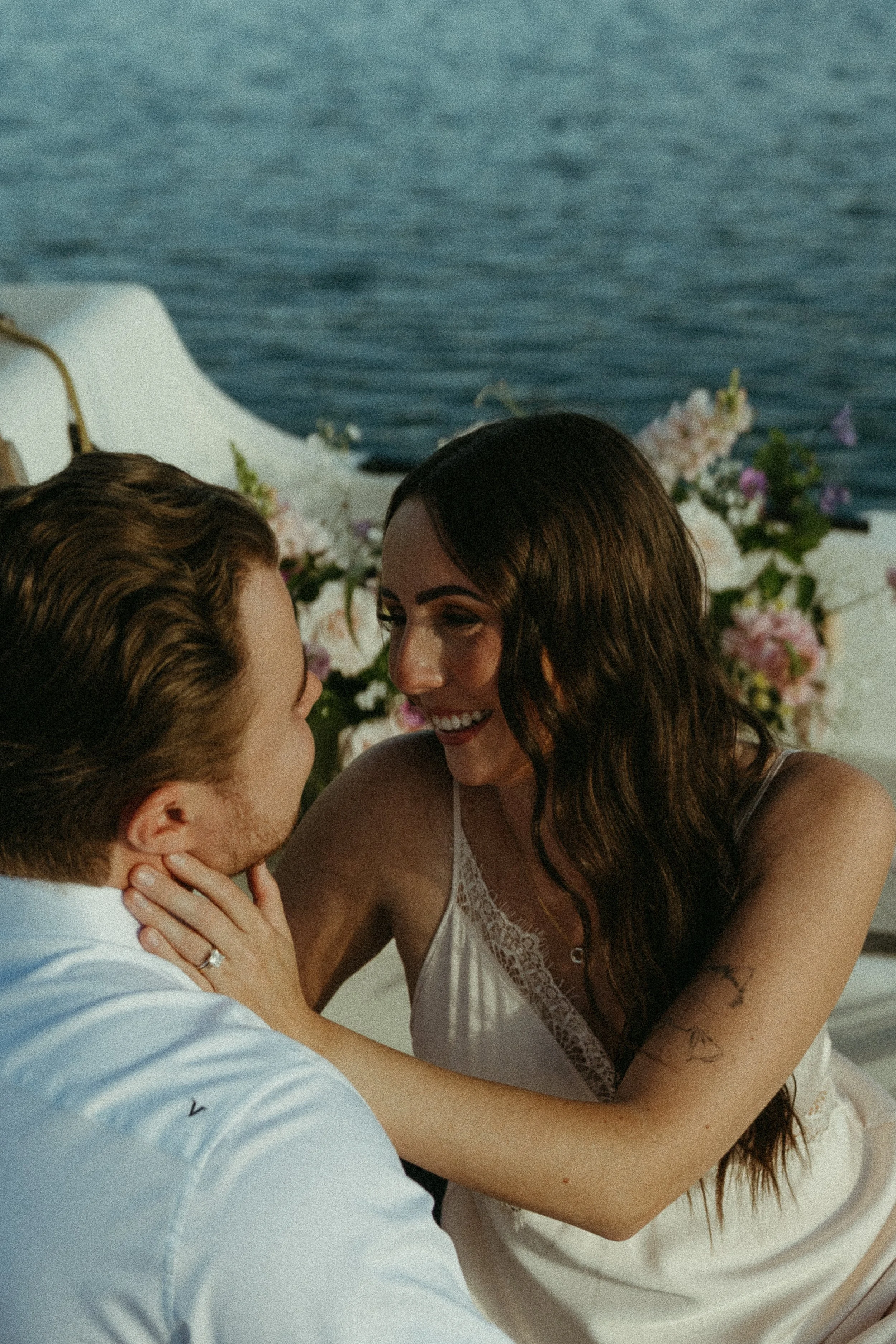 A bride touches the cheek of her groom and smiles into his eyes while they are sat on a sailboat with flowers. August 2025 Elopement/Wedding Sailboat/Sailing Session with KC Photography