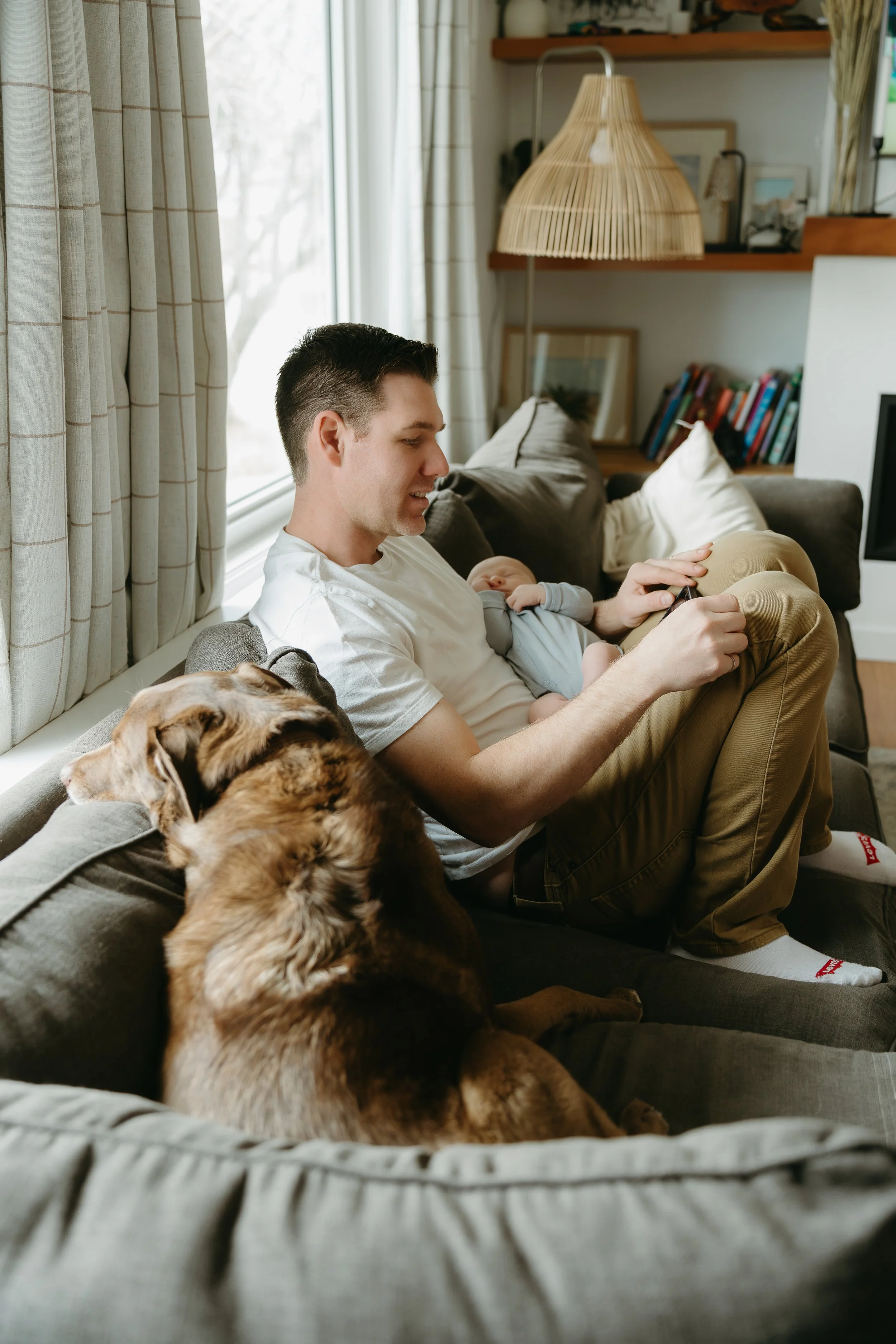 A father sits on the couch in his home holding his newborn baby while his dog sits beside them looking out the window. Calgary Home Lifestyle/Family/Newborn Session with KC Photography