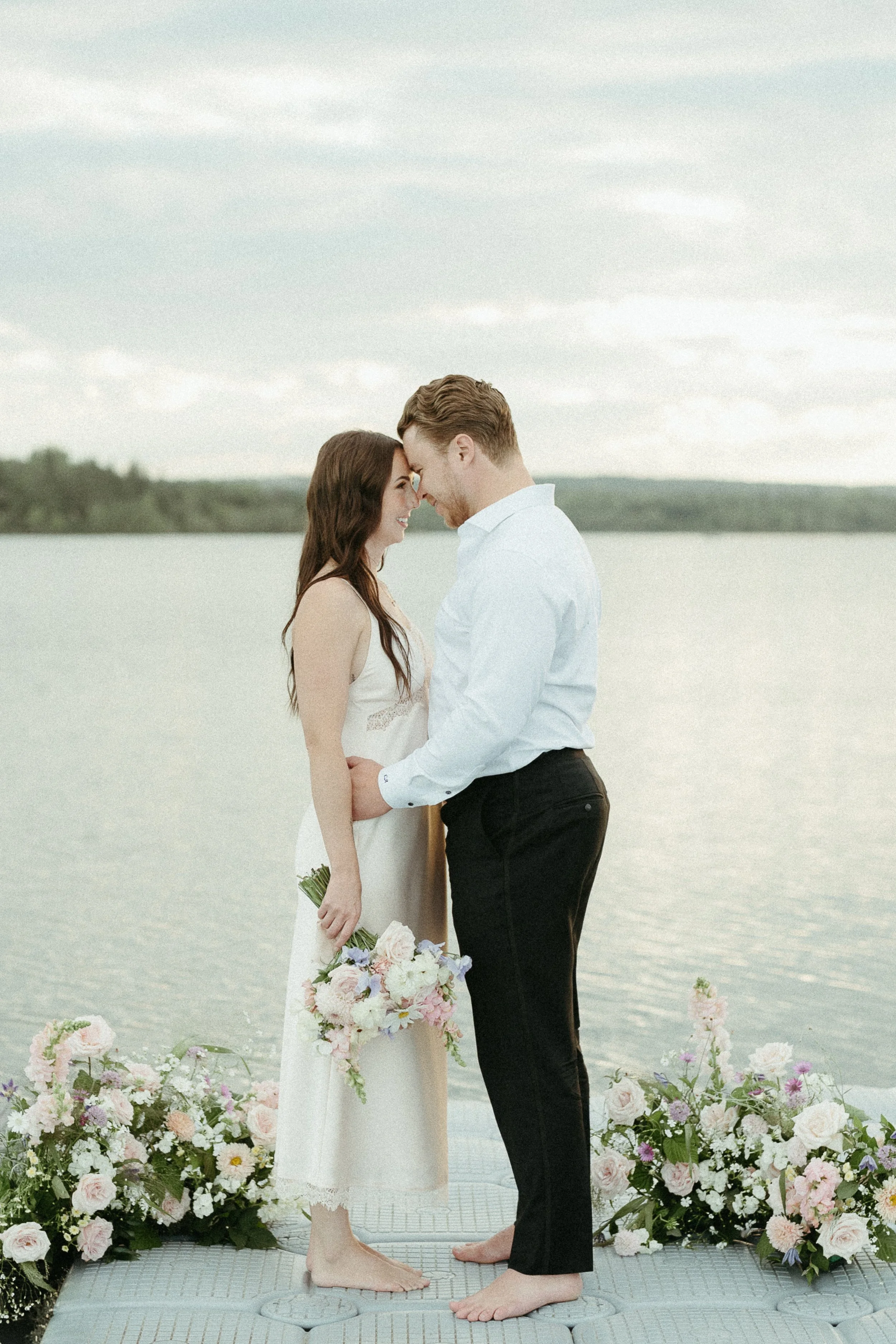 A bride and groom stand touching foreheads and smiling at one another on the end of a dock surrounded by bouquets of flowers and a lake. August 2025 Elopement/Wedding Sailboat/Sailing Session with KC Photography