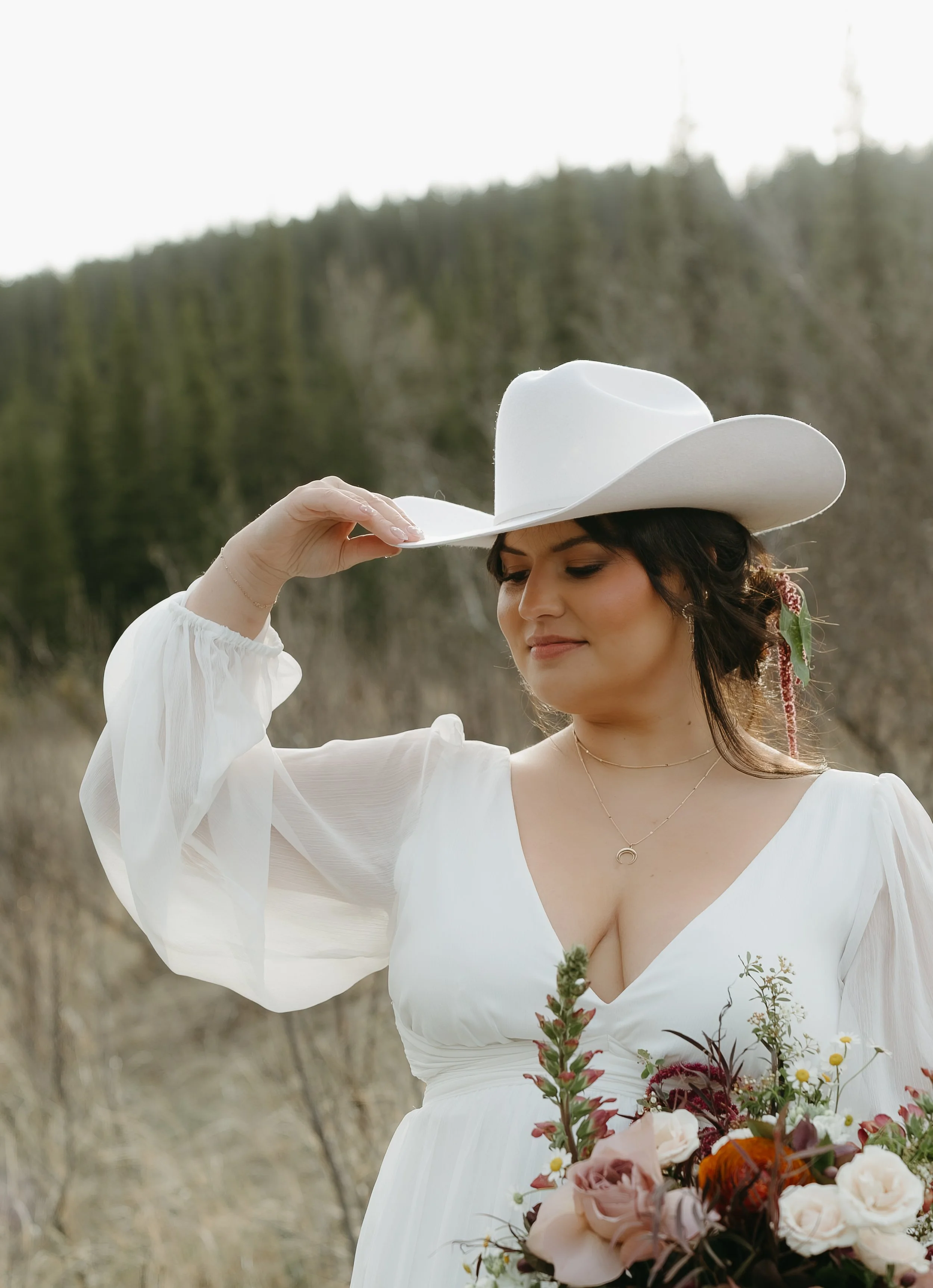 A bride touches her cowboy hat while looking down as she holds a bouquet of flowers in a Kananaskis field surrounded by pine trees. June 2025 Kananaskis Elopement/Wedding Editorial Session with KC Photography