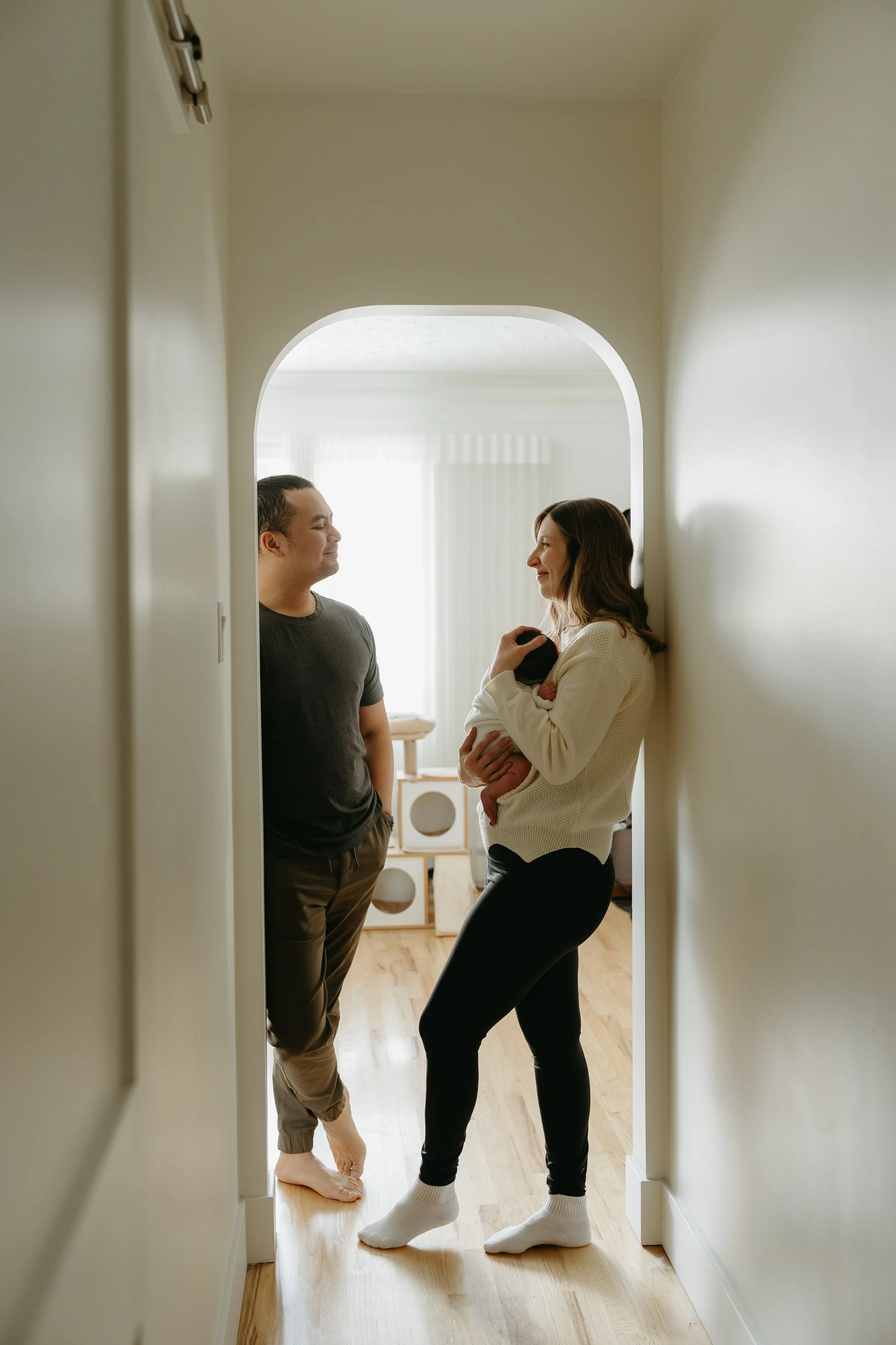 A couple stands in the doorway of their home smiling at each other as the mother holds their newborn baby. Calgary Home Lifestyle/Family/Newborn Session with KC Photography