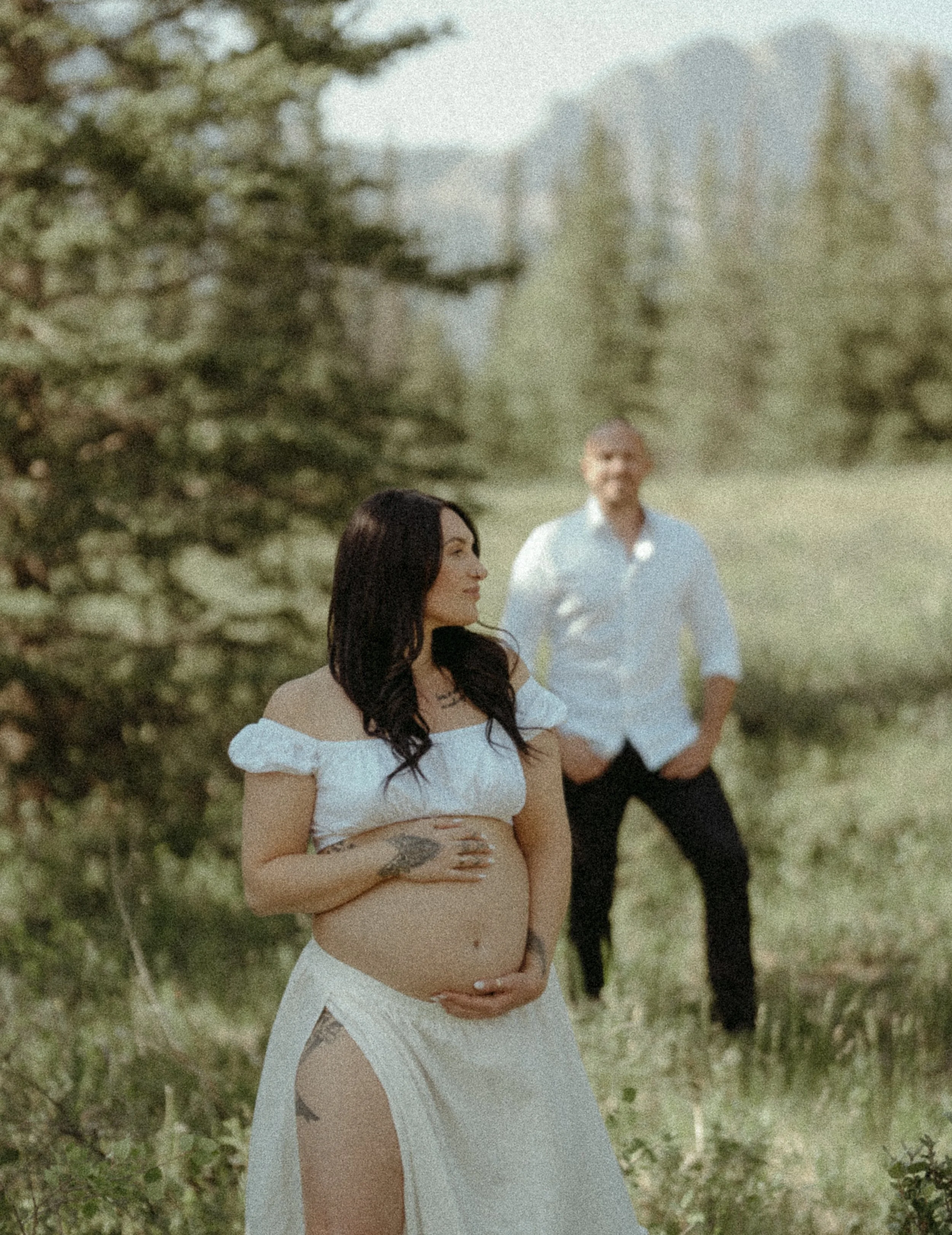 A woman holds her pregnant belly as she looks off into the distance of a Kananaskis field while her husbands looks at her in the background. Kananaskis Lifestyle/Family/Couple/Pregnancy/Maternity Session with KC Photography