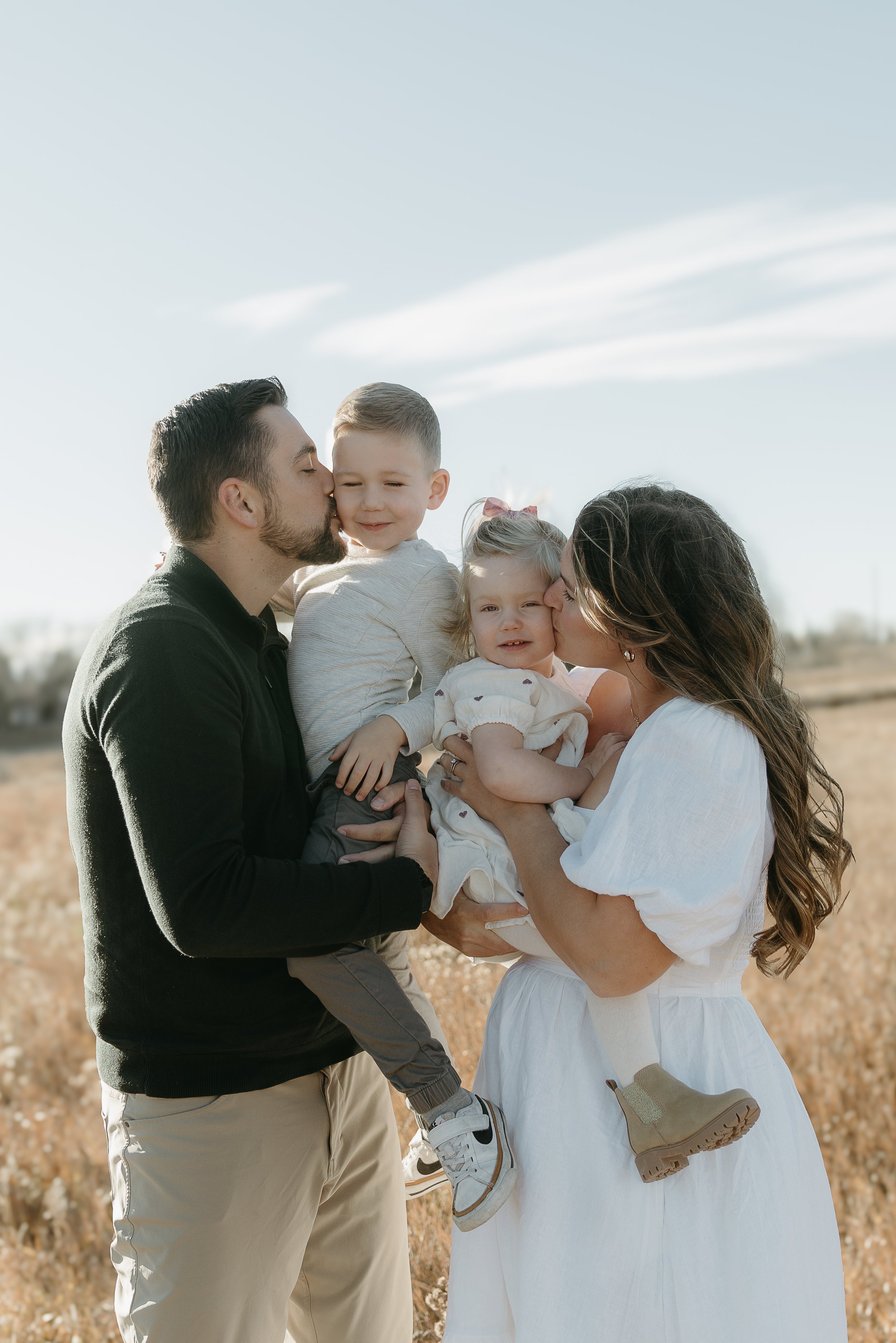 A couple holds their two toddlers between them and each kiss them on the cheek in a field. Calgary Lifestyle/Family Session with KC Photography