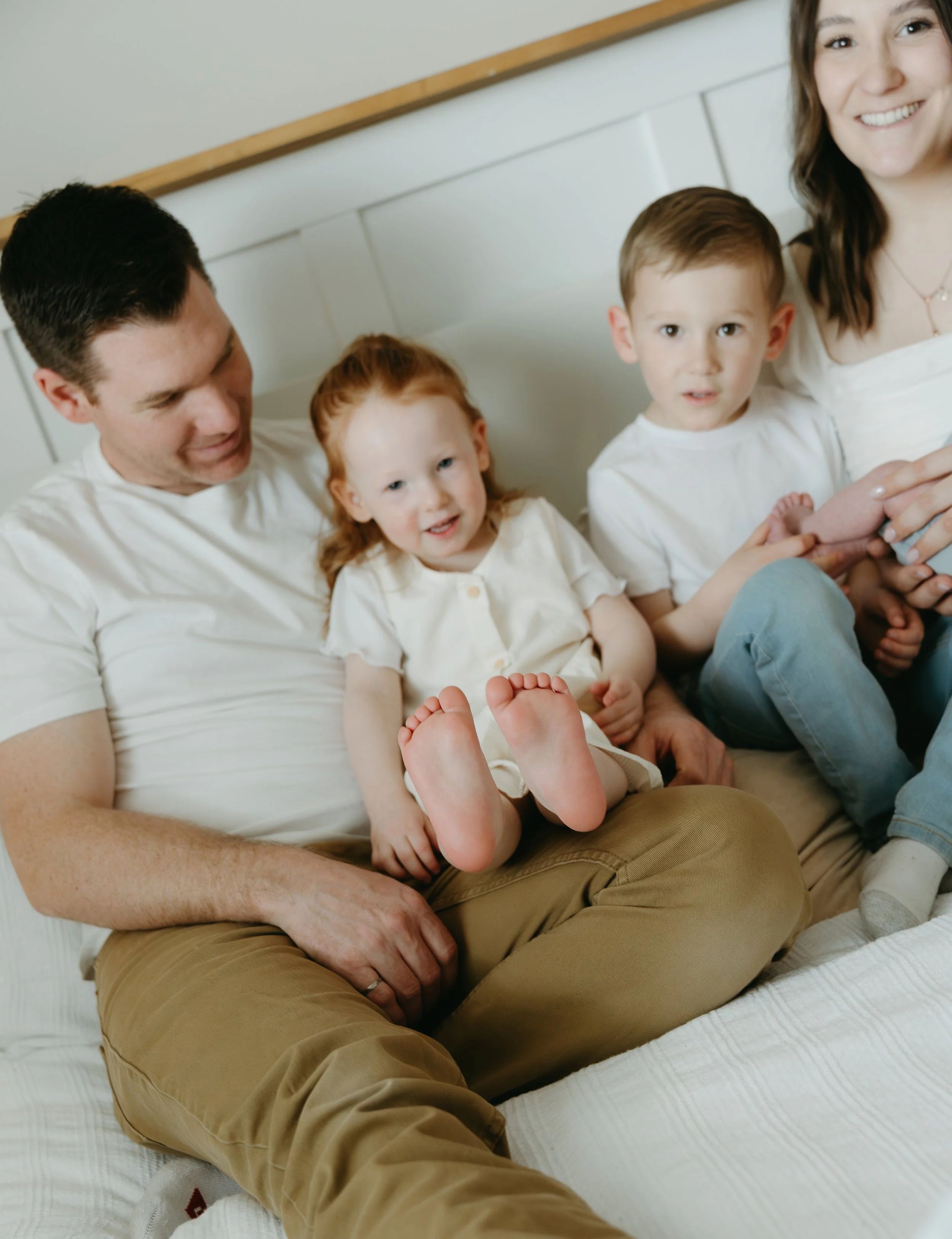 A couple sits on their bed with their two toddlers between them who stick out their bare feet. Calgary Home Lifestyle/Family Session with KC Photography