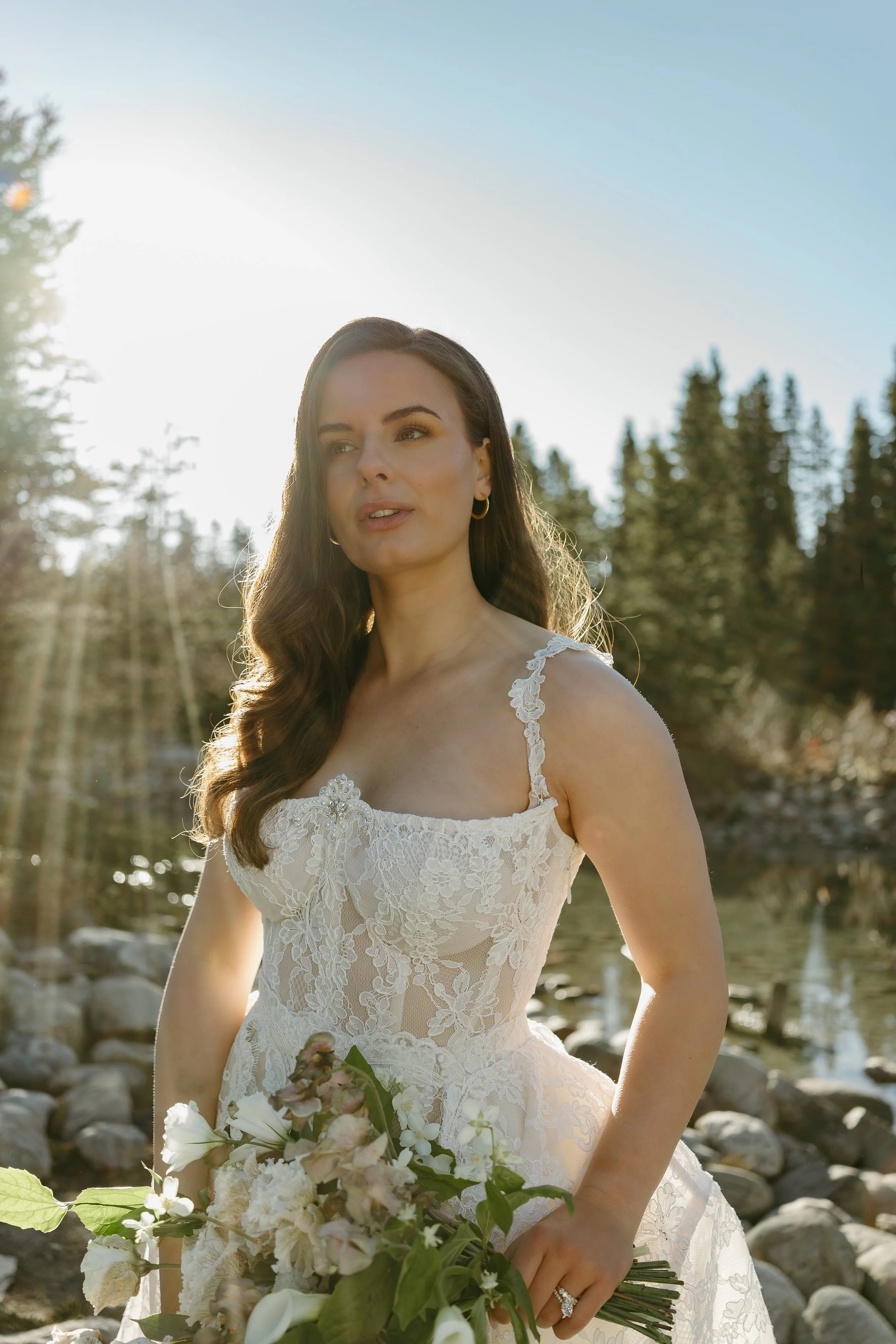 A bride stands on the edge of a mountain creek holding a bouquet of flowers as she looks off into the distance. June 2025 Lake Louise Elopement/Wedding Editorial Session with KC Photography