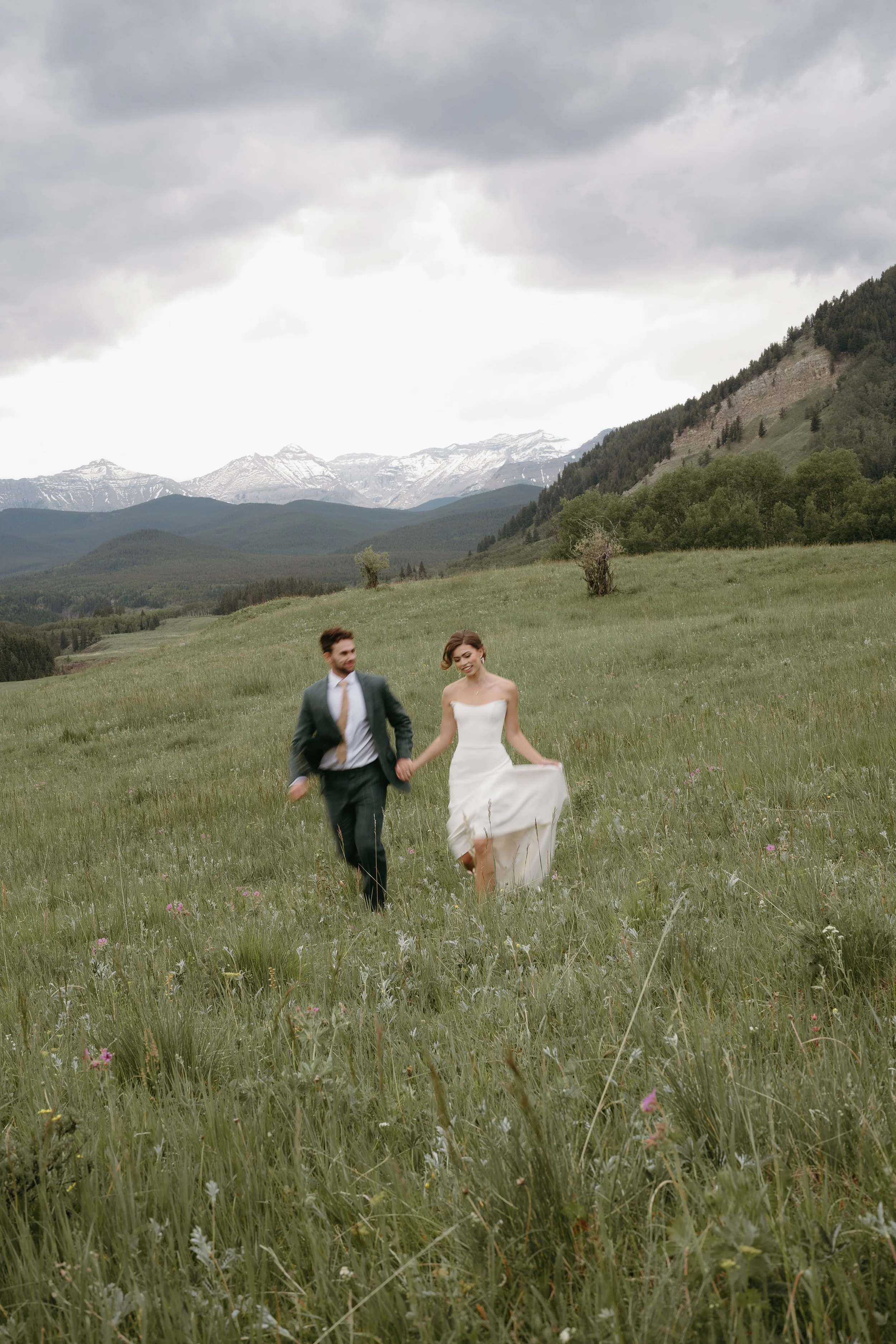 A bride and a groom run through a field holding hands surrounded by mountains. June 2025 Kananaskis Elopement/Wedding Session with KC Photography