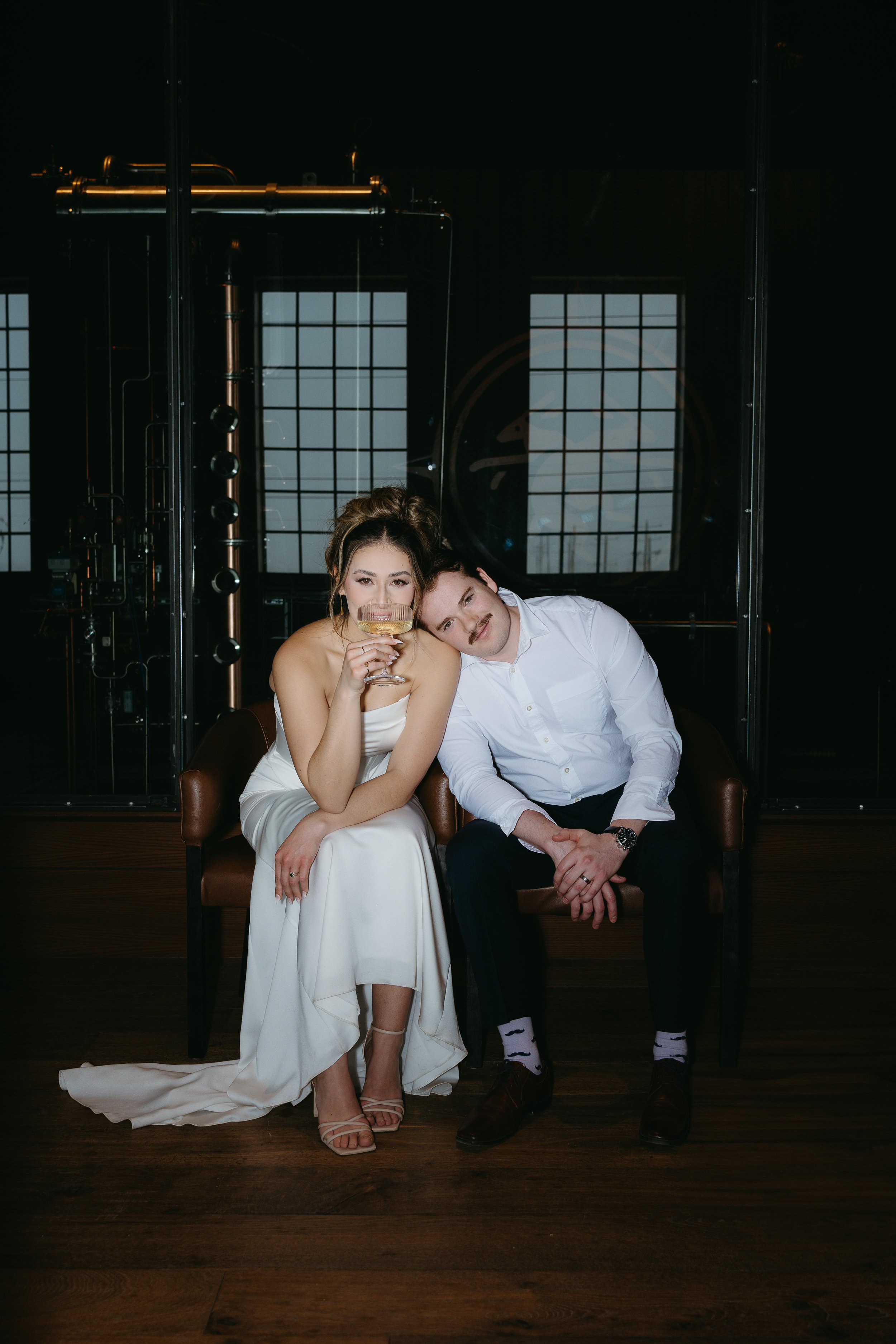 A groom sits beside his bride on a bench while resting his head on her shoulder while she sips a glass of champagne. Calgary Elopement/Wedding Session with KC Photography