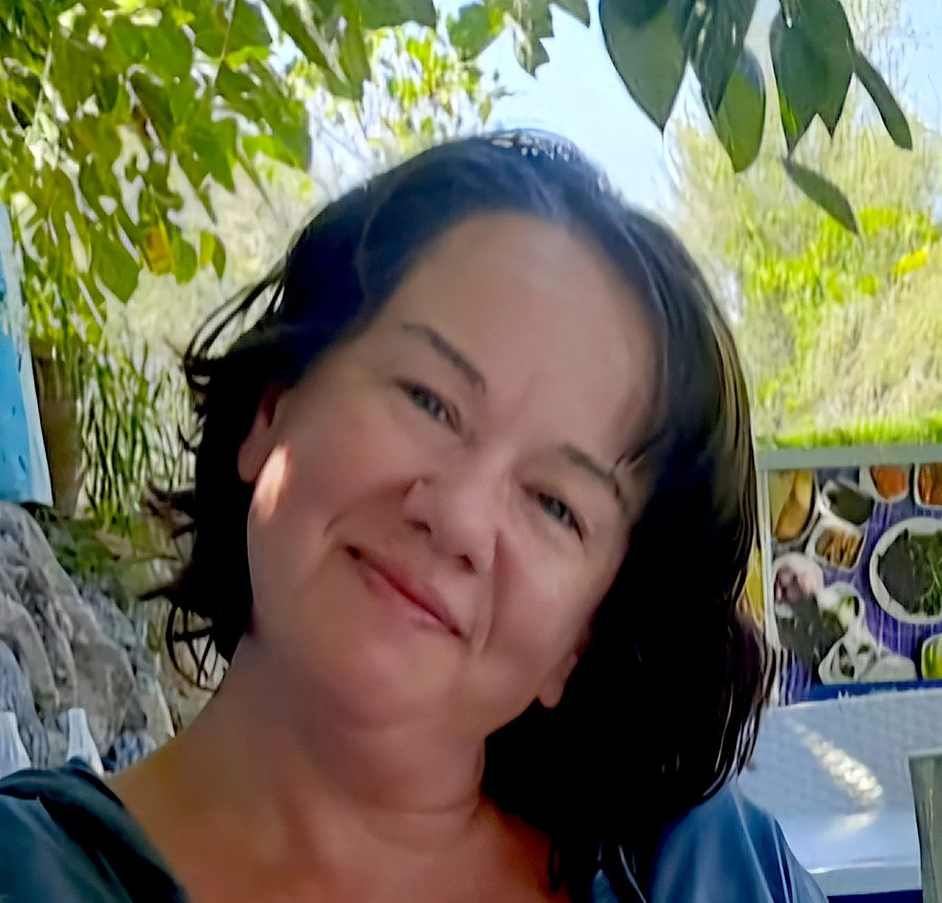 A woman with black hair smiling outdoors with trees and foliage in the background.
