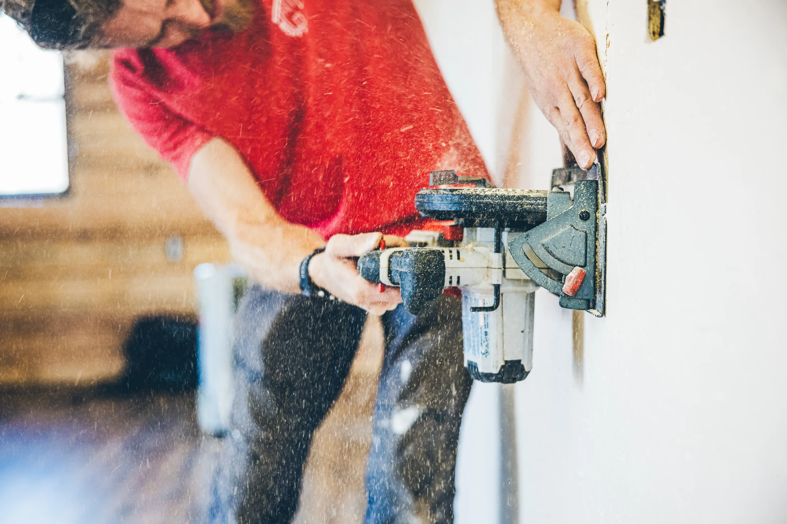 A person wearing a red shirt using a power tool on a white wall, with sawdust visible in the air.