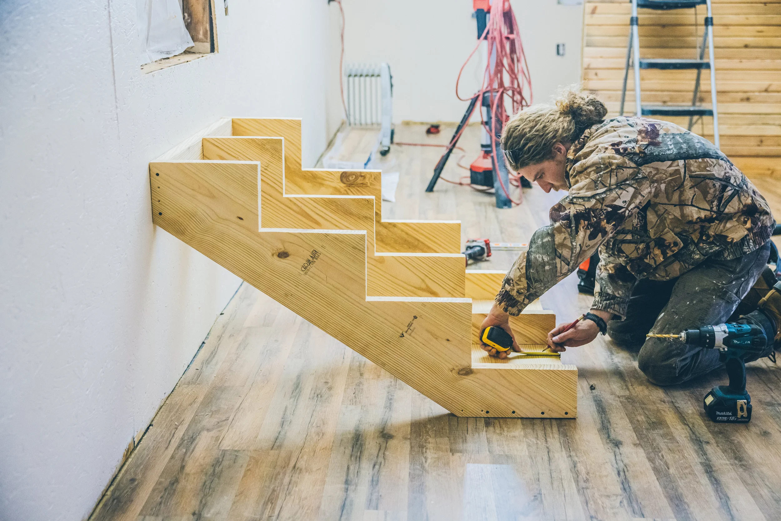 A person in camouflage clothing working on a wooden staircase indoors, using a measuring tape and surrounded by tools, with construction materials and equipment in the background.
