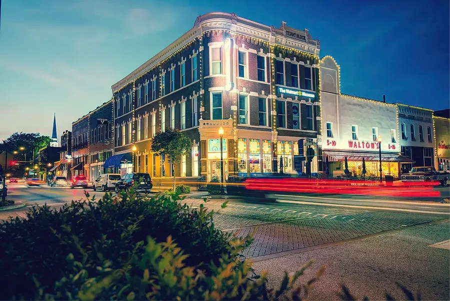 Night view of a historic downtown Bentonville, Arkansas decorated with string lights, with cars and street activity in the foreground.