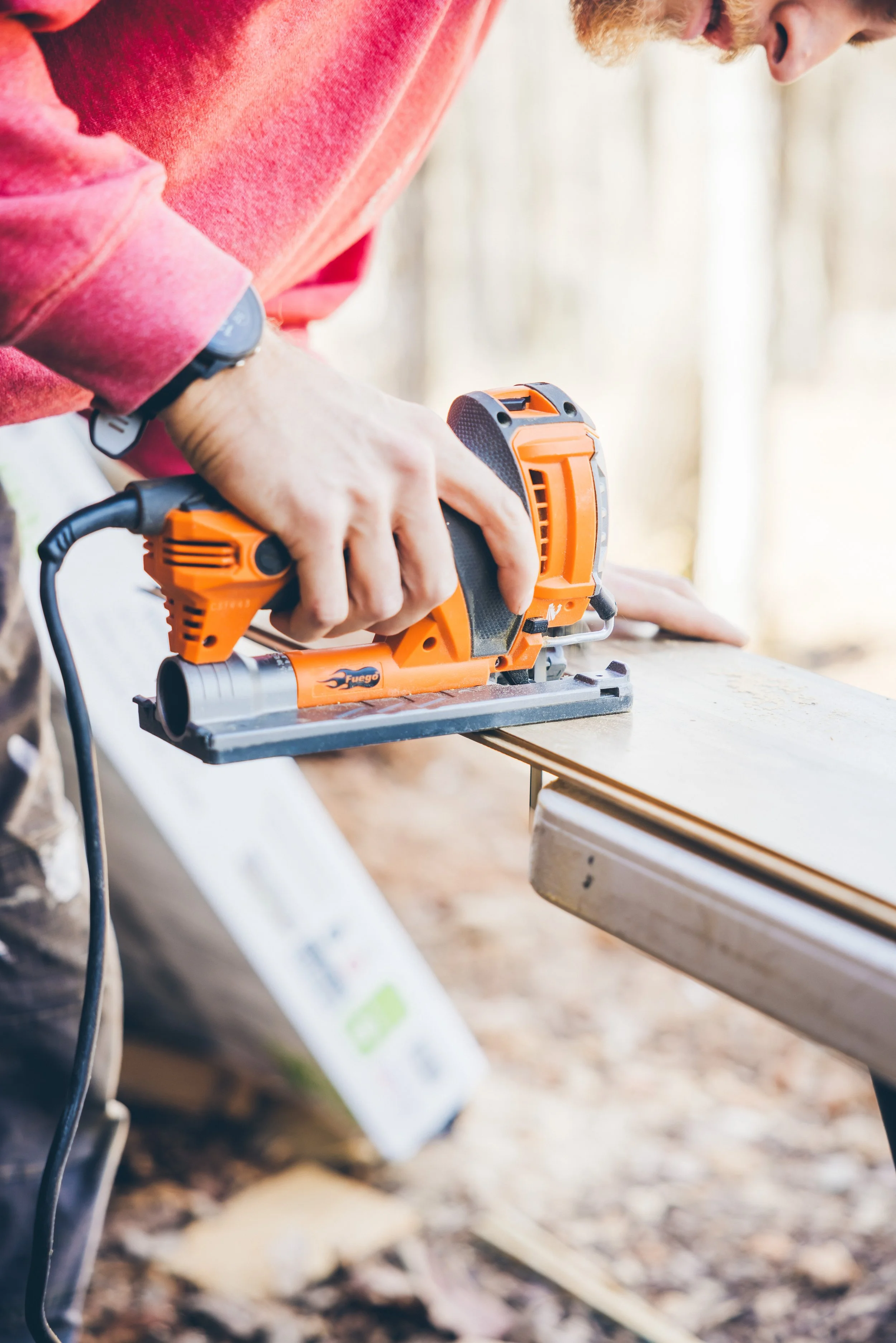A person wearing a red shirt and a black wristwatch uses an orange power sander to sand a wooden surface.