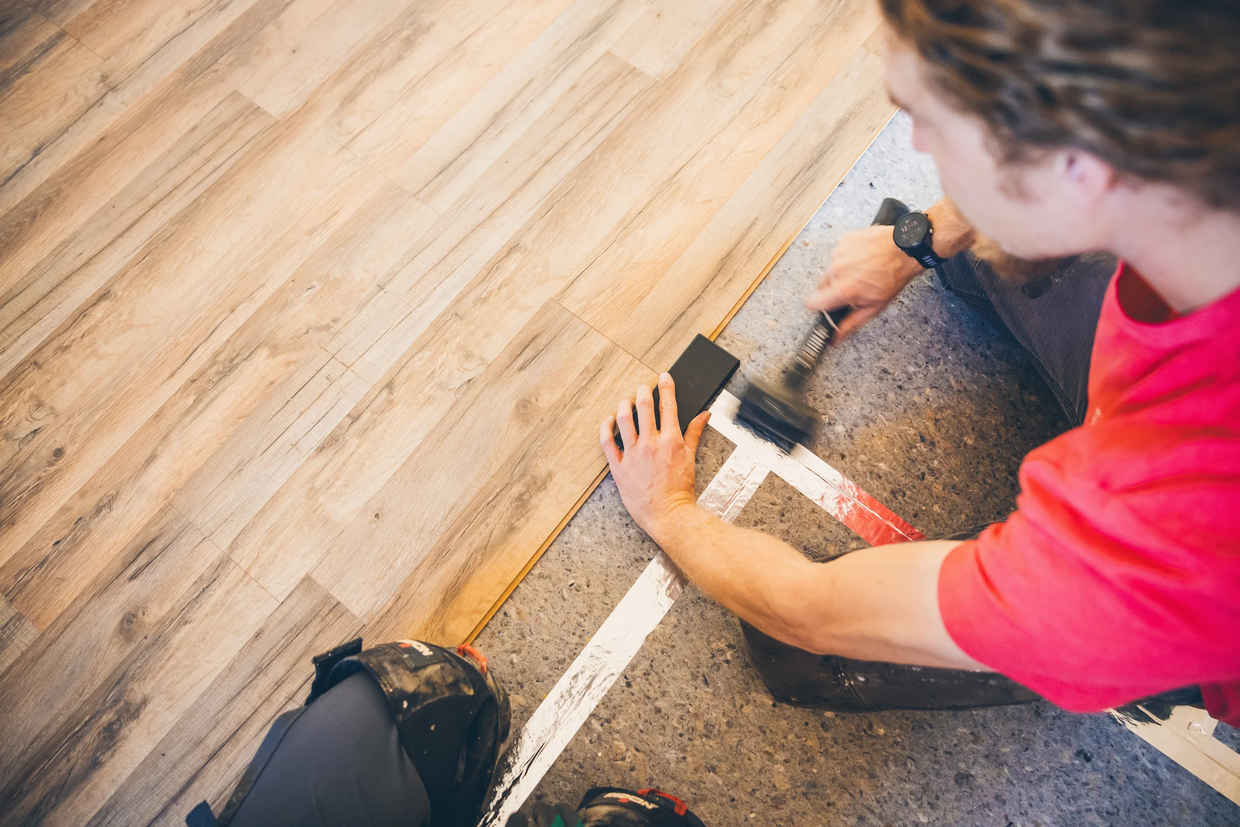 Person installing hardwood flooring, aligning a plank with a measuring tool, wearing a red shirt, black pants, and a wristwatch, with a pencil and tools nearby.
