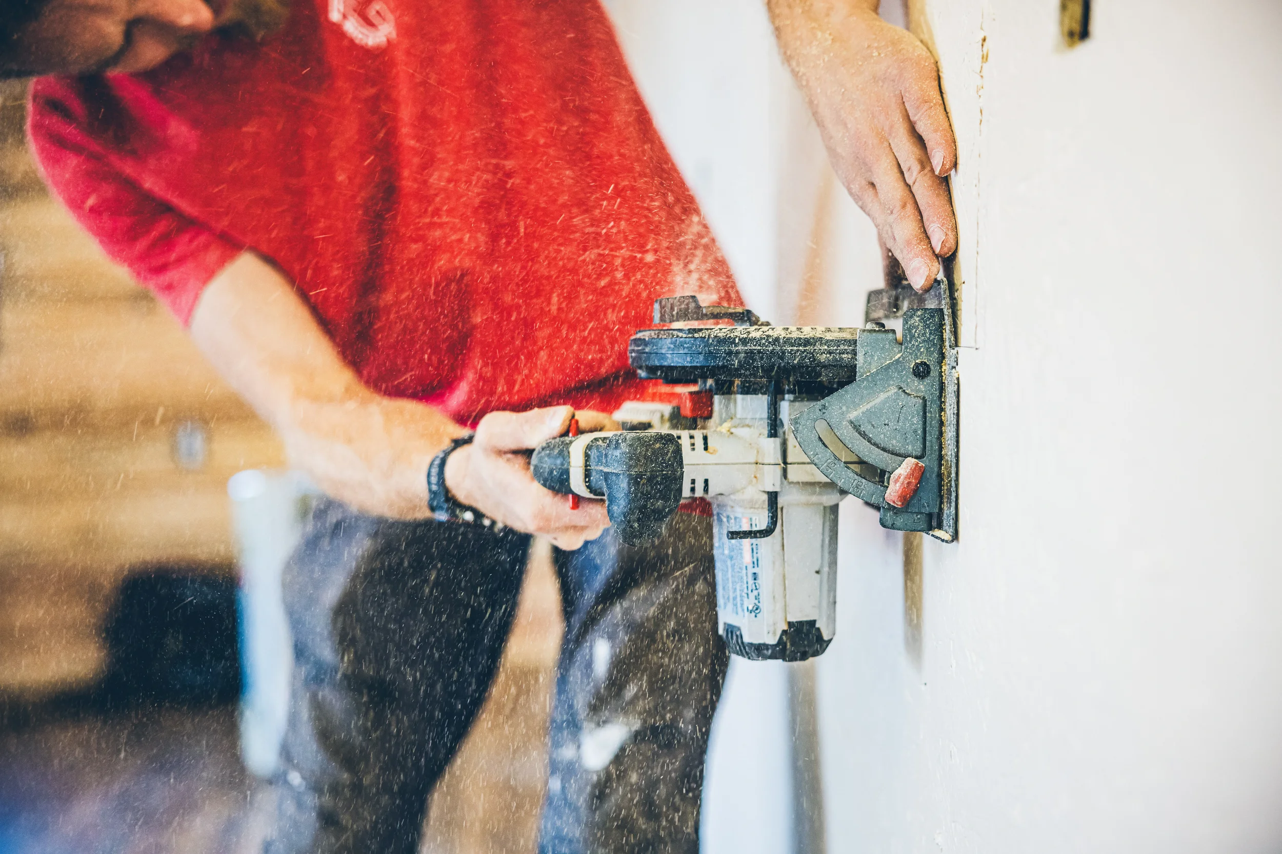 Person in a red shirt using a power drill on a wall, with dust flying around.