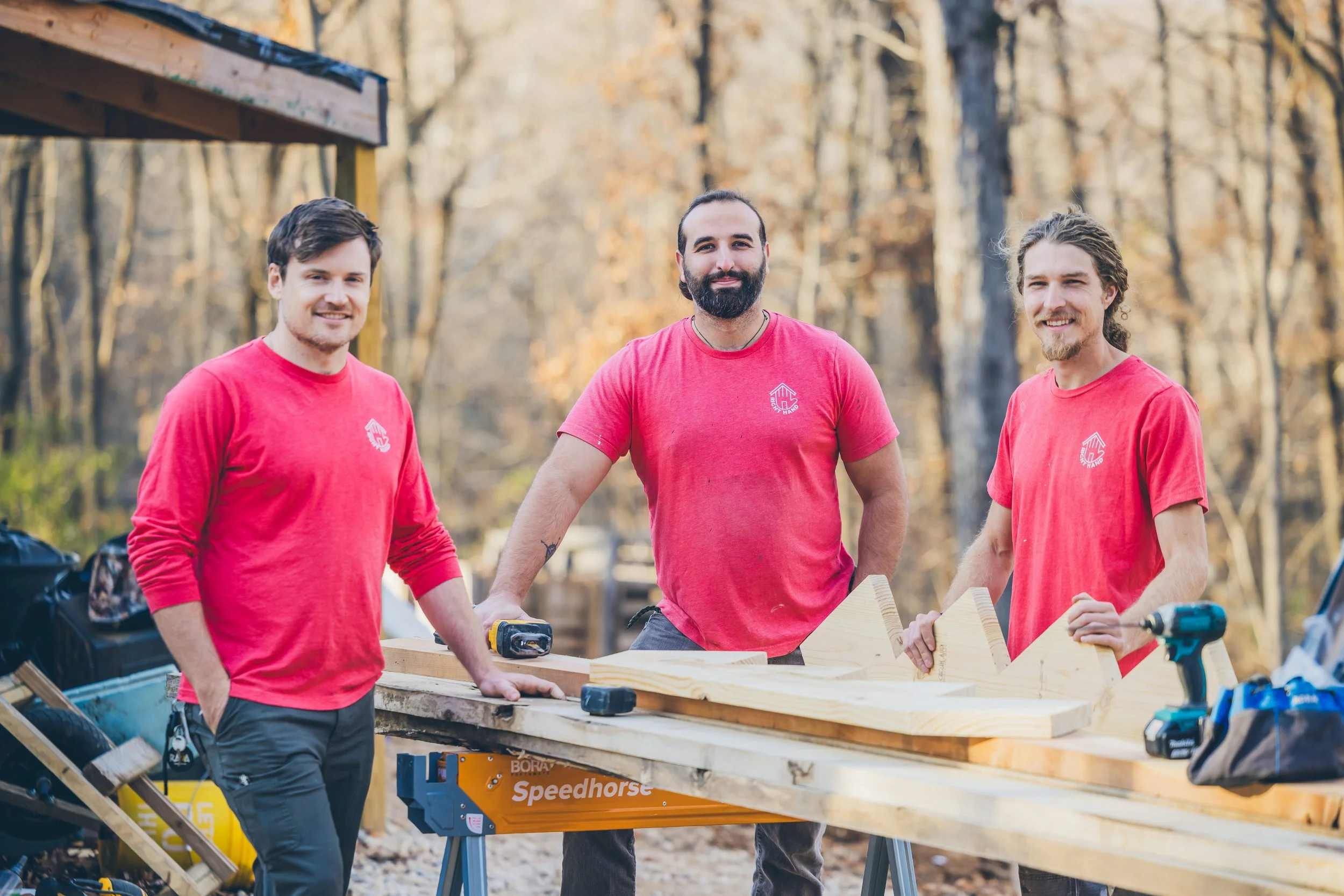 Three men wearing red shirts working outdoors on a woodworking project in a wooded area during autumn.