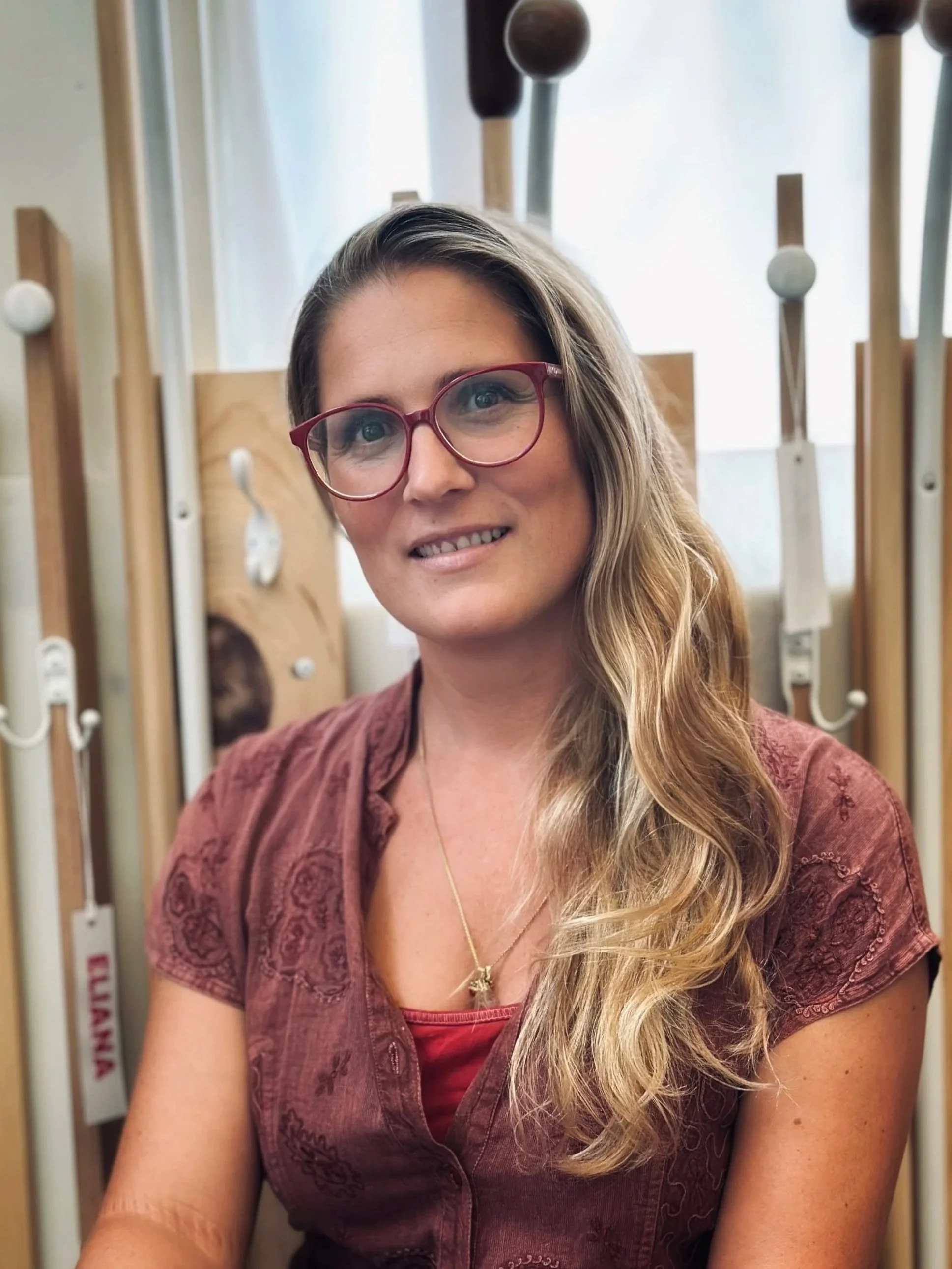 A woman with long wavy blonde hair wearing red glasses and a maroon blouse, smiling.