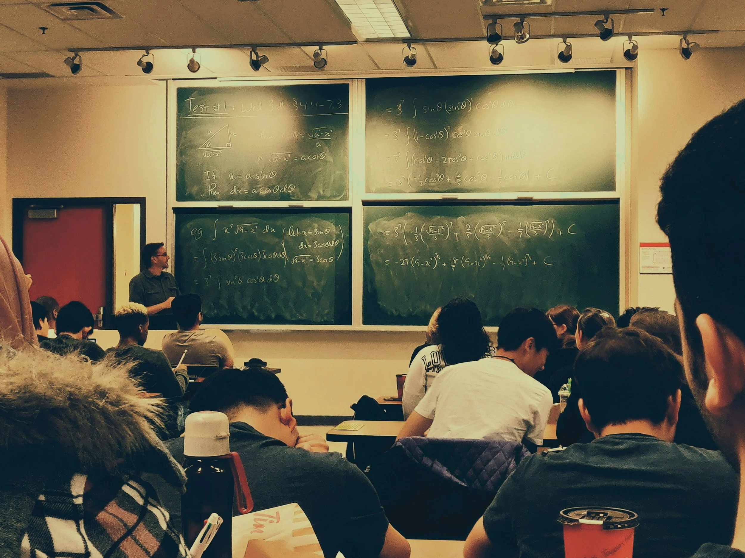 College classroom with students seated at desks, some taking notes, listening to a professor standing next to green chalkboards filled with mathematical equations.