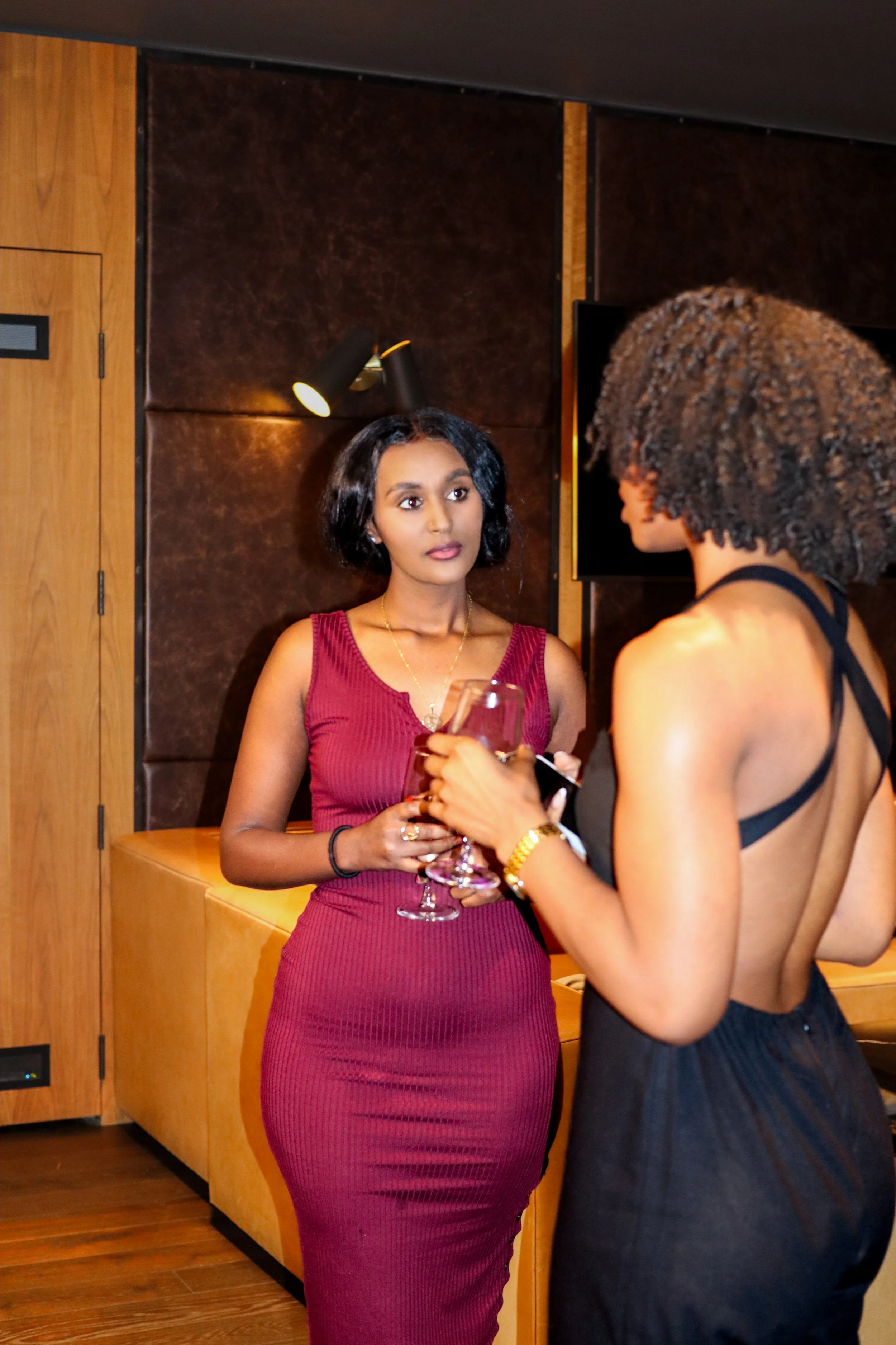 Two women conversing indoors, one in a sleeveless burgundy dress holding a wine glass, the other in a black backless dress.