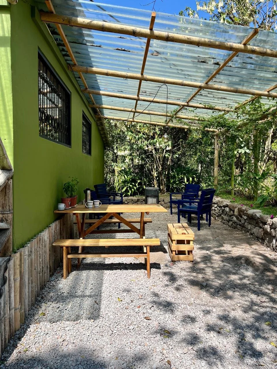 An outdoor patio area with a green wall on the left, three black chairs on the right, a picnic table with benches on the left, and a stone-bordered garden bed on the right. There is a transparent corrugated roof supported by bamboo poles overhead and