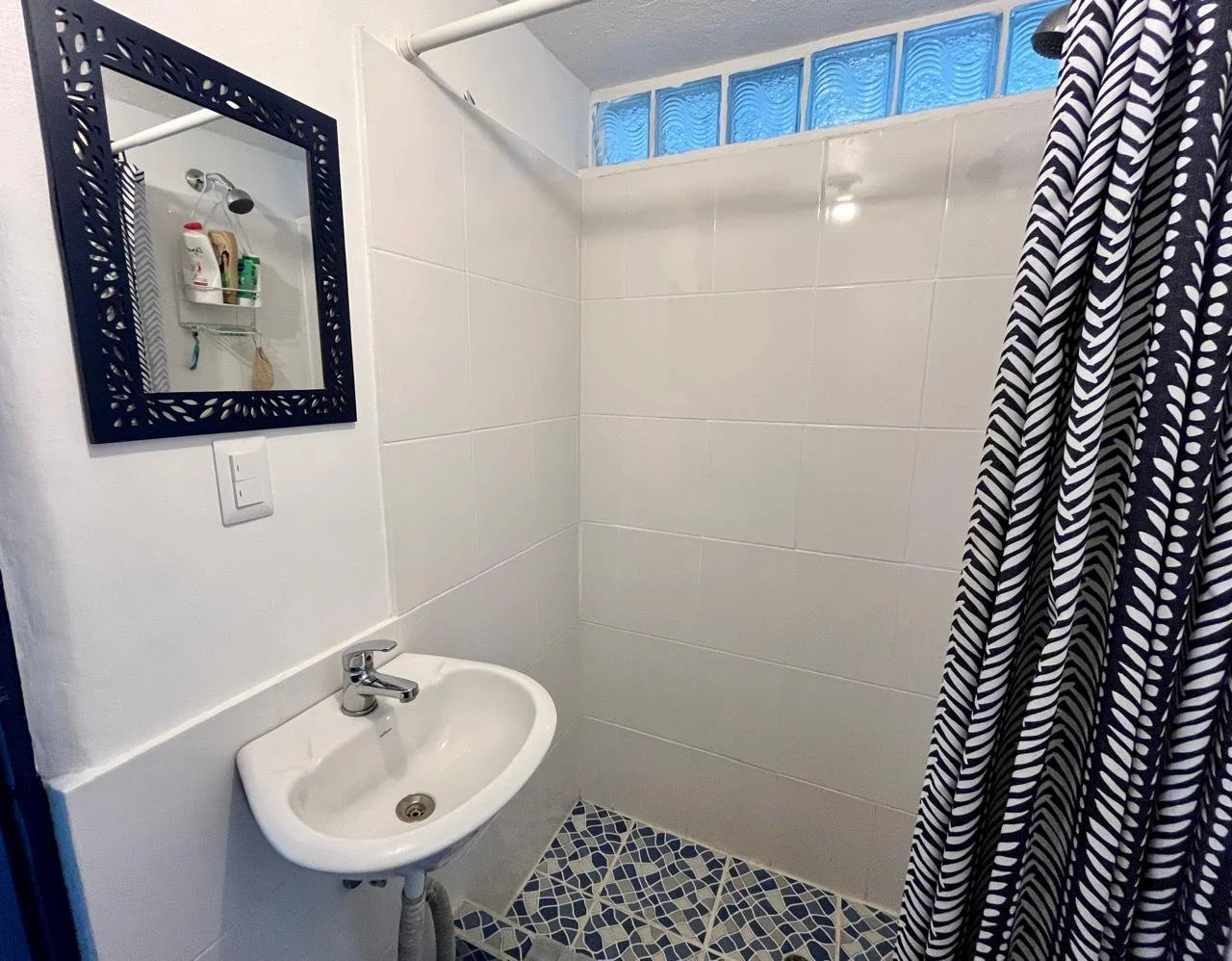 Small bathroom with a white sink, a black-framed mirror, a shower area with a striped blue and white shower curtain, and a row of glass blocks near the ceiling.