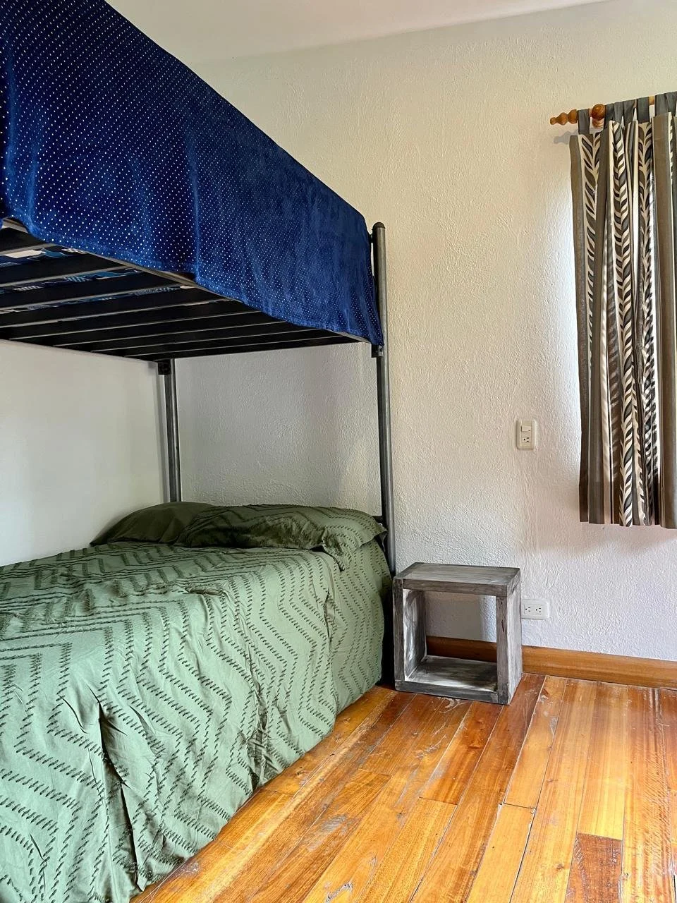 A room with wooden floor, a bed with green cover, a bunk bed with blue bedding, a small gray wooden side table, a white wall, a window with patterned curtain, and an electrical outlet.