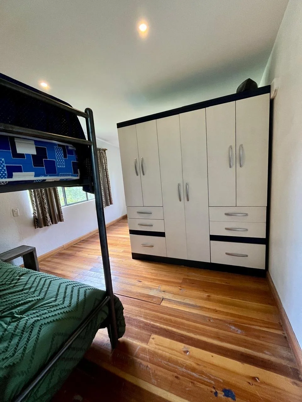 A bedroom with wood flooring, a white wardrobe, a bunk bed with a green mattress, and a window with curtains.