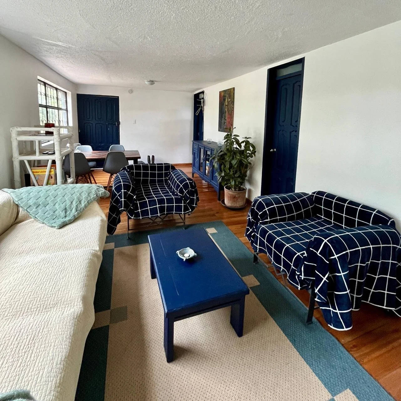 Living room with white couch, two blue checkered armchairs, a blue coffee table, and a blue sideboard. There is a potted plant, a dining table with chairs, and blue doors. The floor is wooden, and the walls are white.