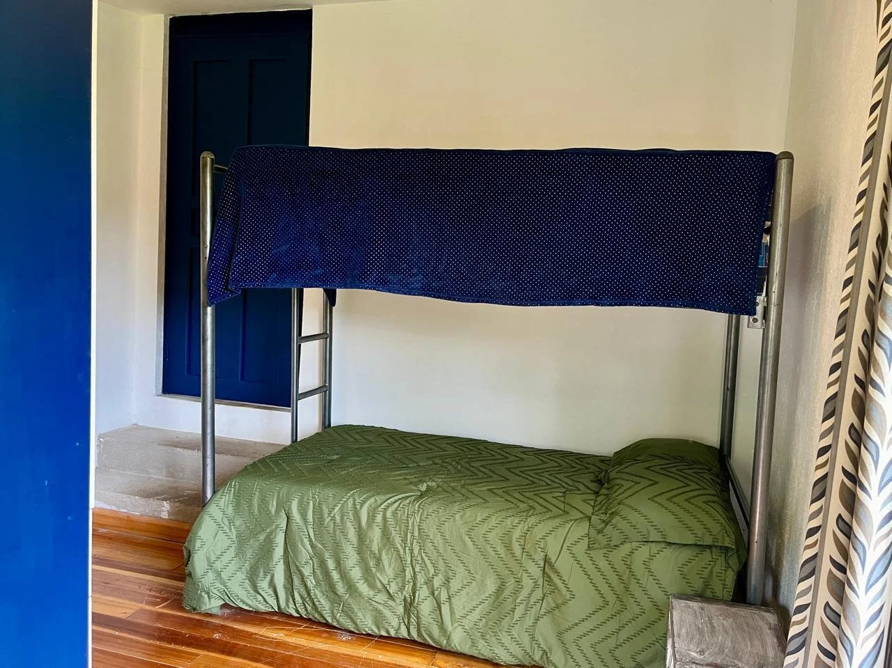 A room with a bunk bed featuring a metal frame, a green bedspread on the bottom bunk, and a blue mattress cover on the top bunk. There is a small wooden table next to the bottom bunk and a dark blue door in the background.