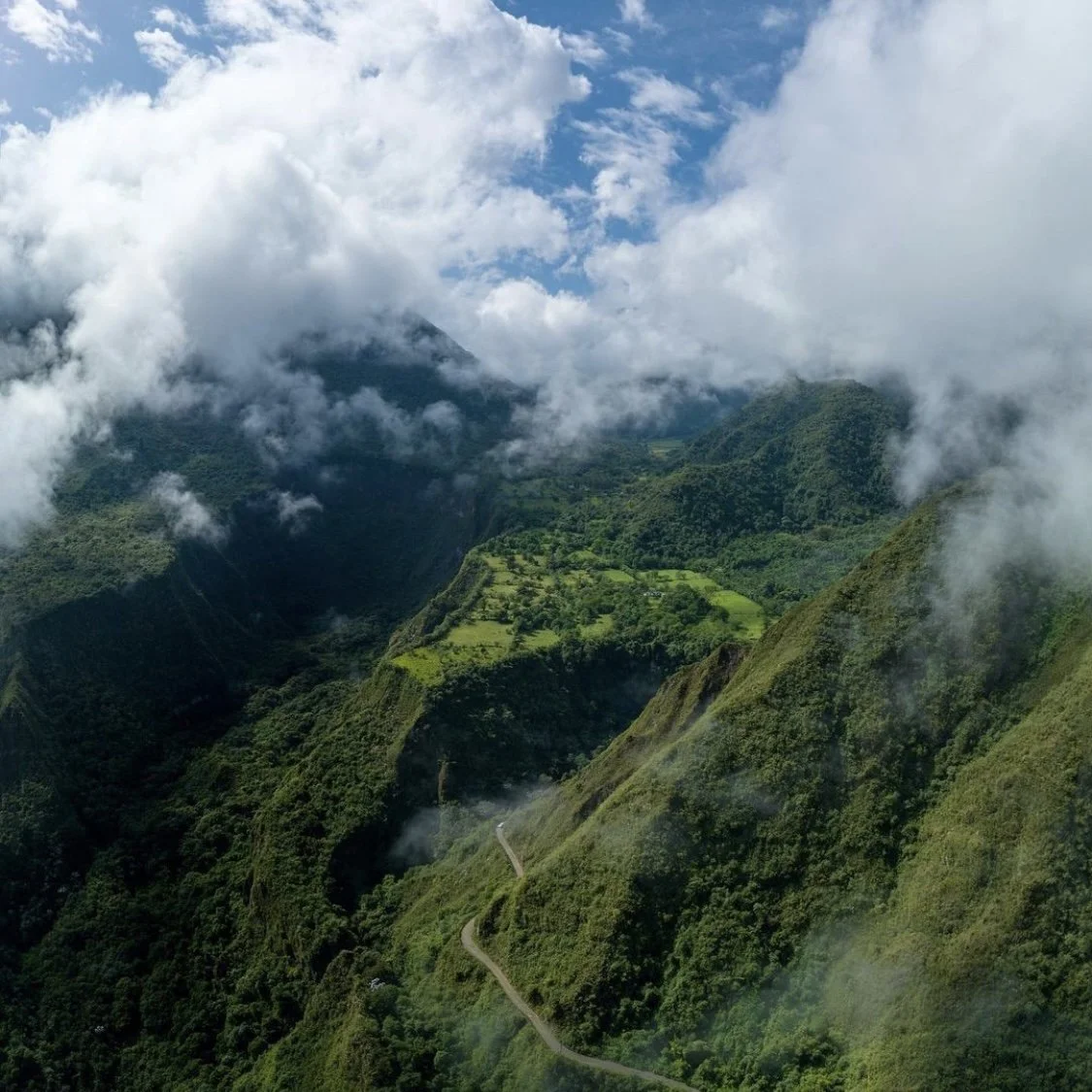 Lush green mountains with a winding road, partly covered by clouds and mist, under a cloudy sky.
