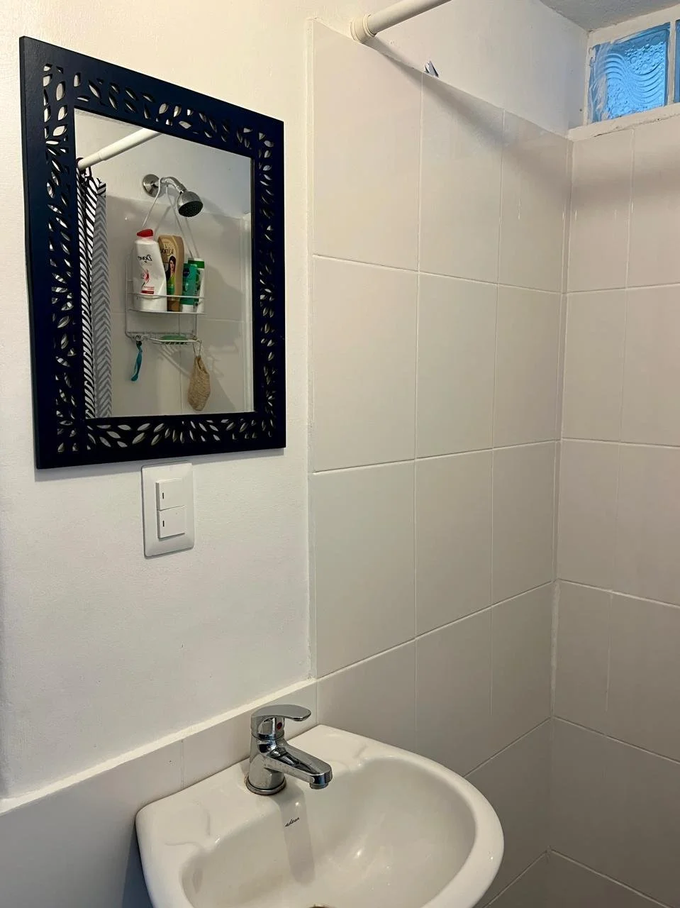 Bathroom corner with a small white sink with a silver faucet, a mirror with a black decorative frame, and a shower area with white tiles and a showerhead. The mirror reflects a shower caddy holding toiletries.