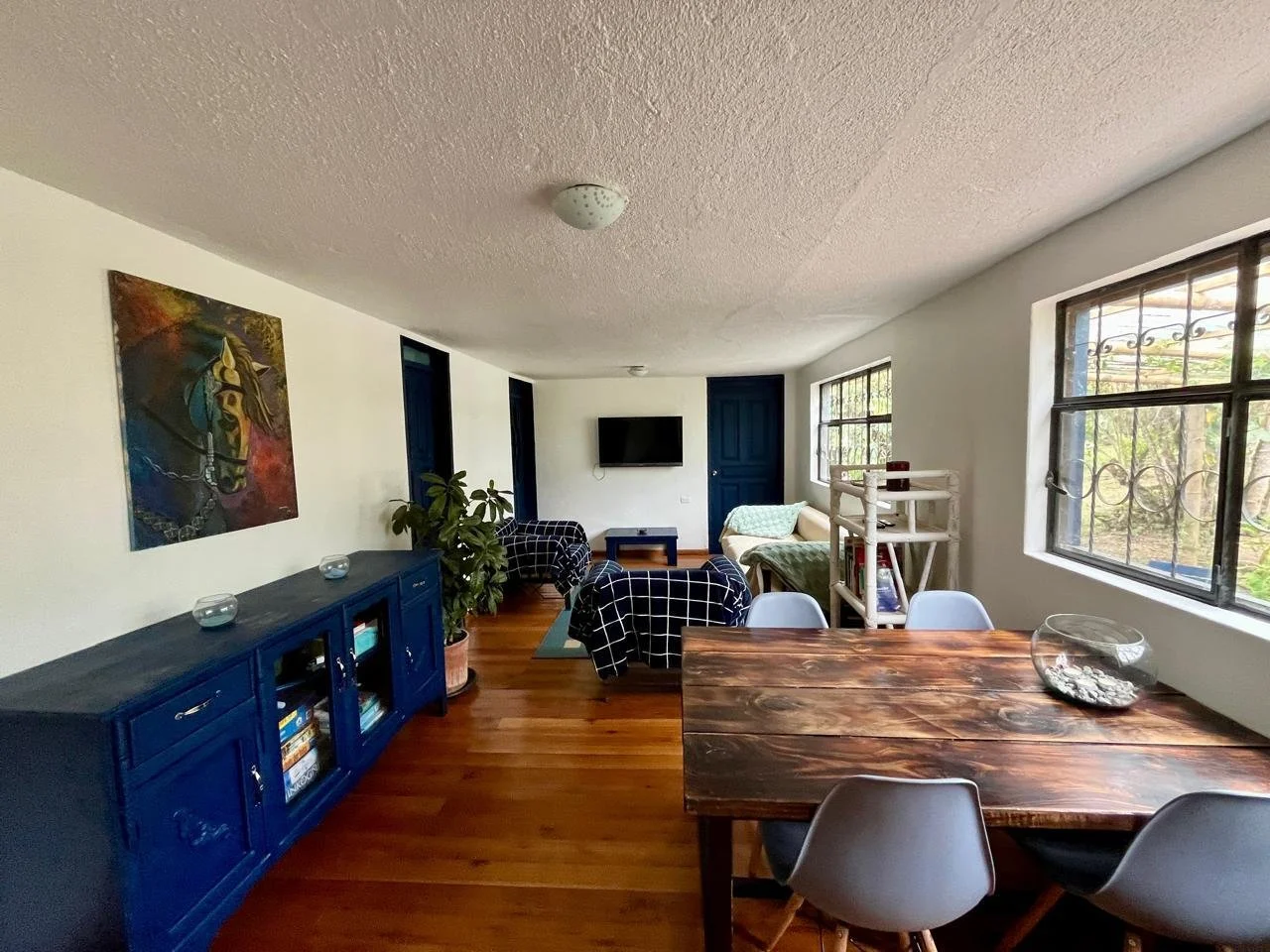 Living room with wooden floors, a blue sideboard, a wall-mounted TV, a large window, a dining table with white chairs, a sitting area with two armchairs and a sofa, and a painting of a horse on the wall.