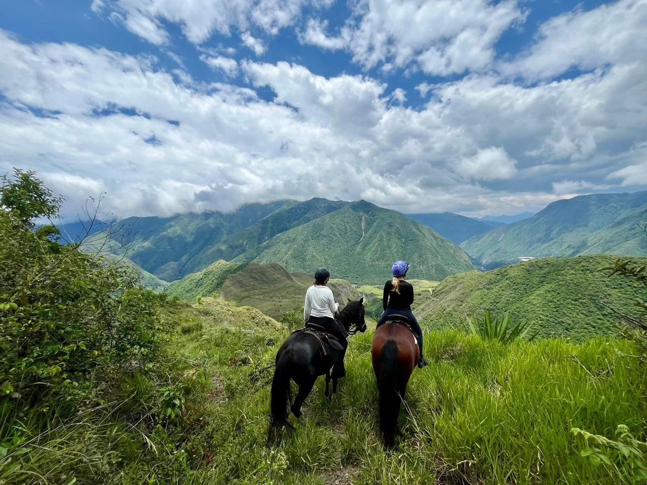 Two people riding horses through green grass with mountainous landscape and partly cloudy sky in the background.