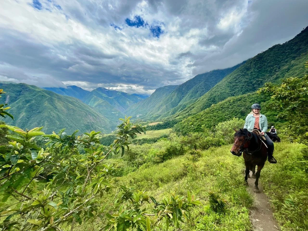 Person riding a horse on a narrow trail through lush green mountains under a partly cloudy sky.