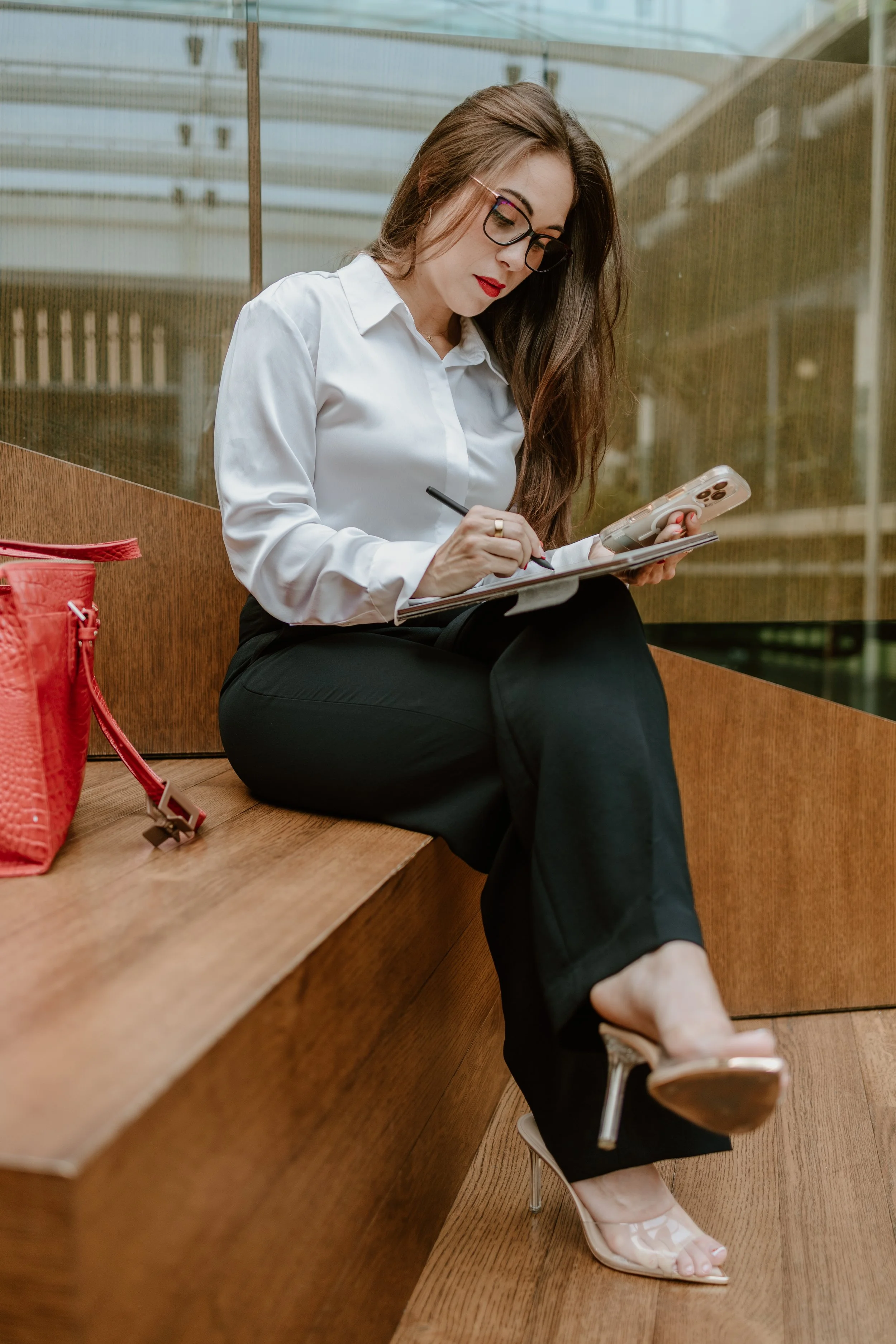 A woman with long brown hair, wearing glasses, red lipstick, a white blouse, black pants, and clear high-heeled shoes, sitting on a wooden bench in a modern indoor space, looking at her phone and writing in a notebook.