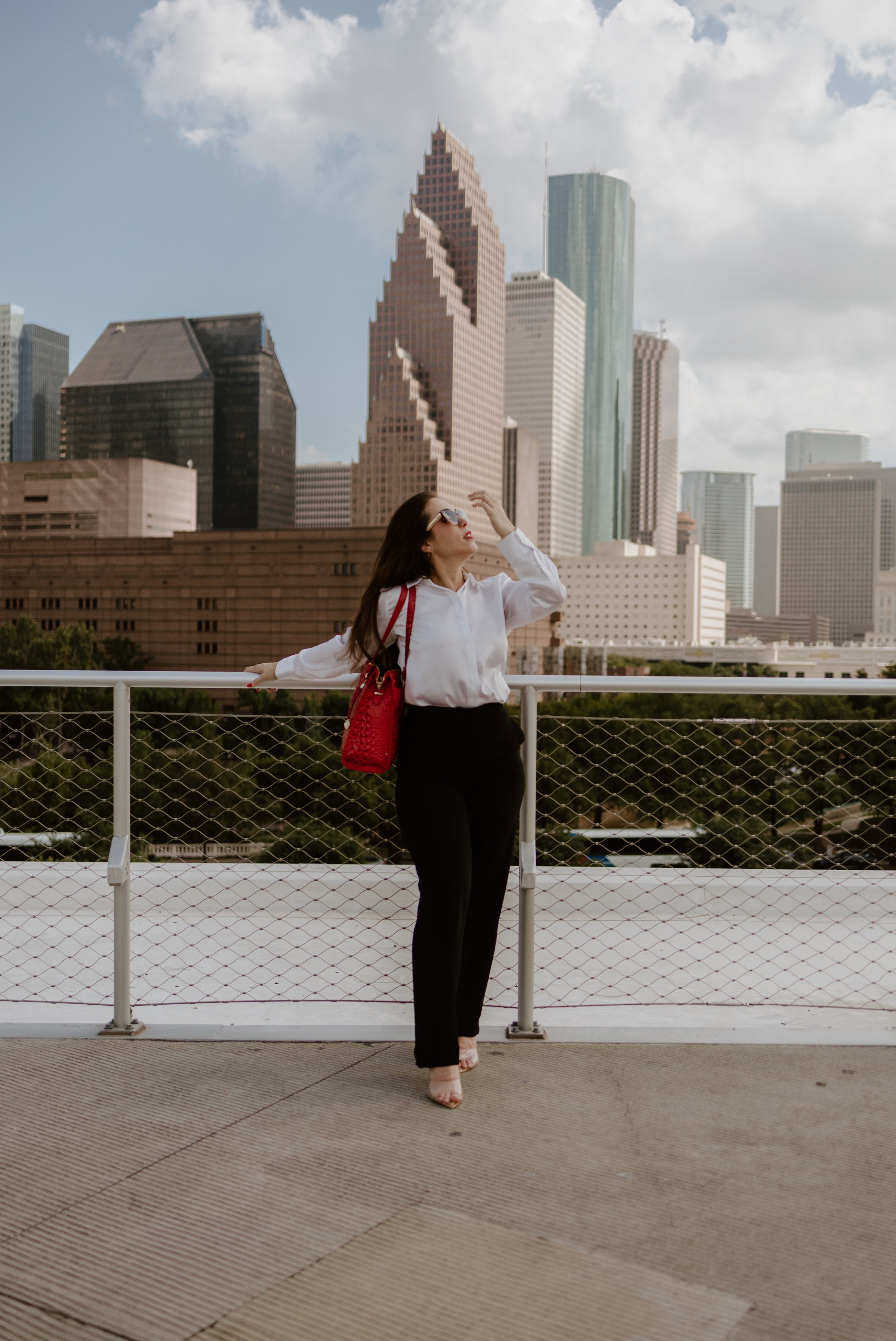 A woman dressed in black pants and a white blouse, carrying a red bag, standing on an outdoor balcony with a city skyline in the background, under a partly cloudy sky.