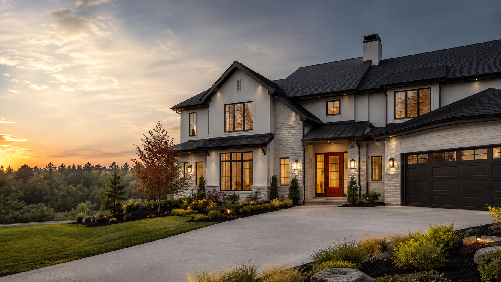 A modern two-story house with a black garage door, white exterior walls, and a stone facade, illuminated by exterior lights at sunset, with landscaped yard and trees surrounding it.
