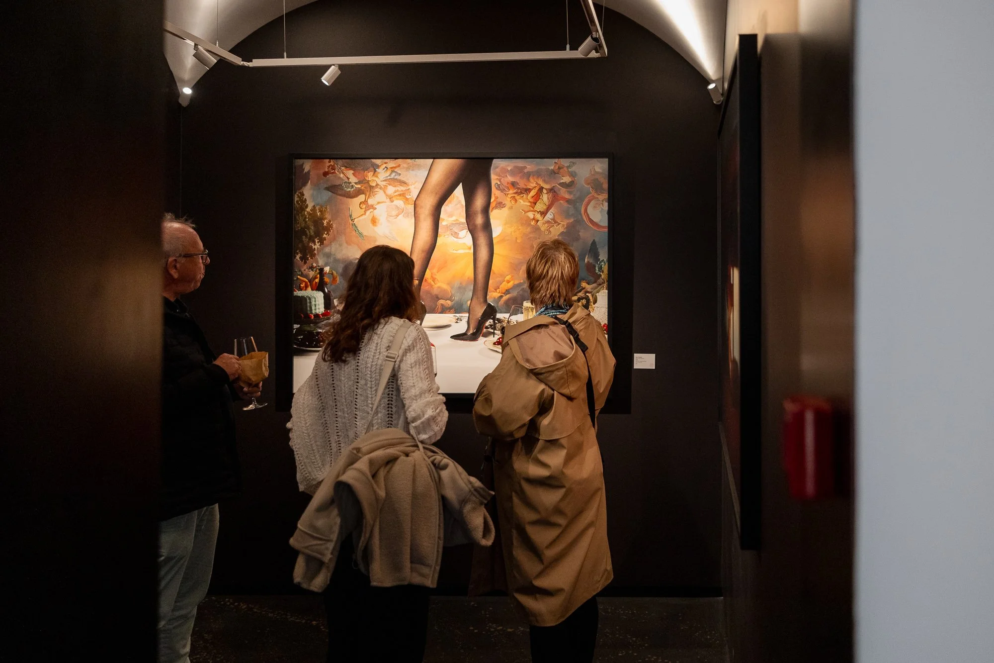 People viewing a large painting of a woman with long legs surrounded by clouds and flying figures in an art gallery.