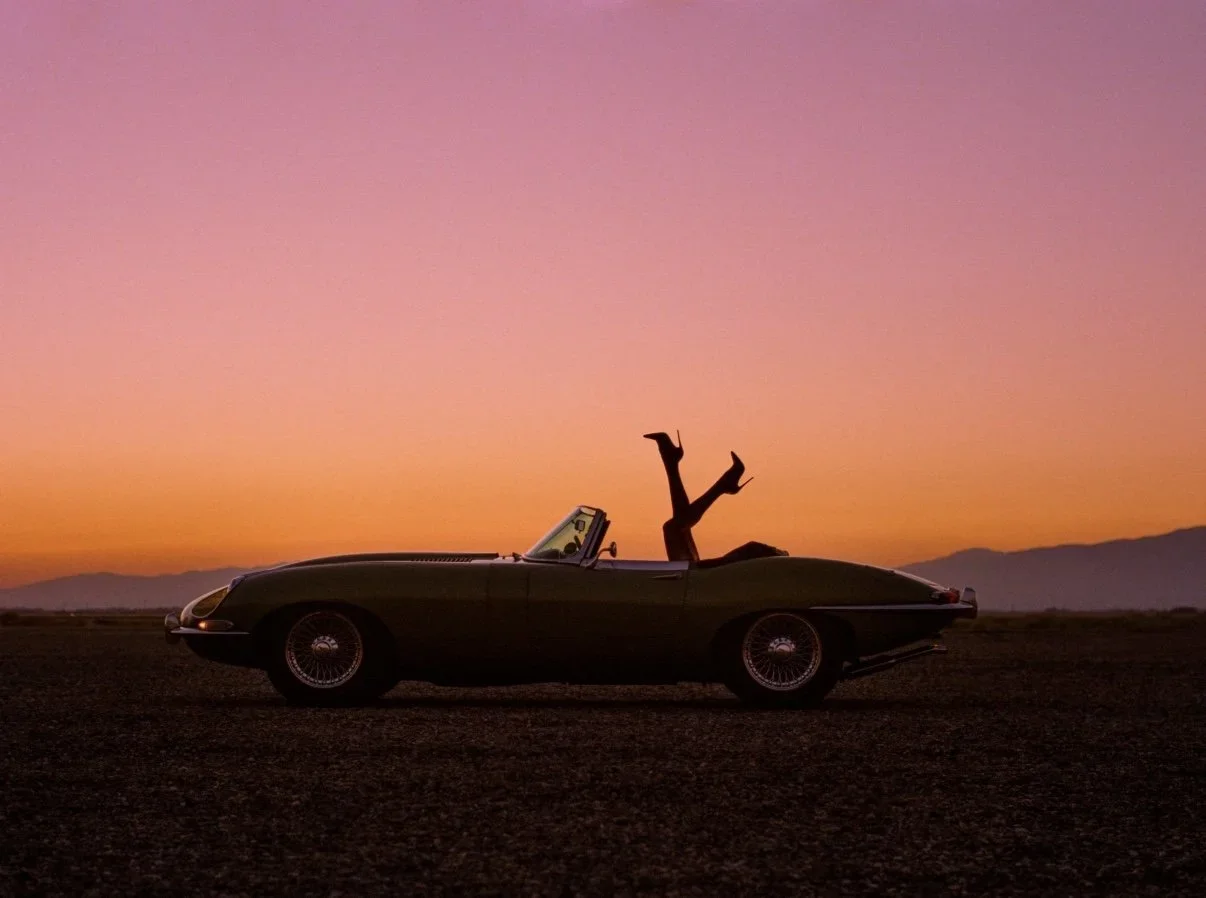 A person doing a handstand on the trunk of a vintage convertible car during sunset or sunrise, with a mountain range in the background and colorful sky.