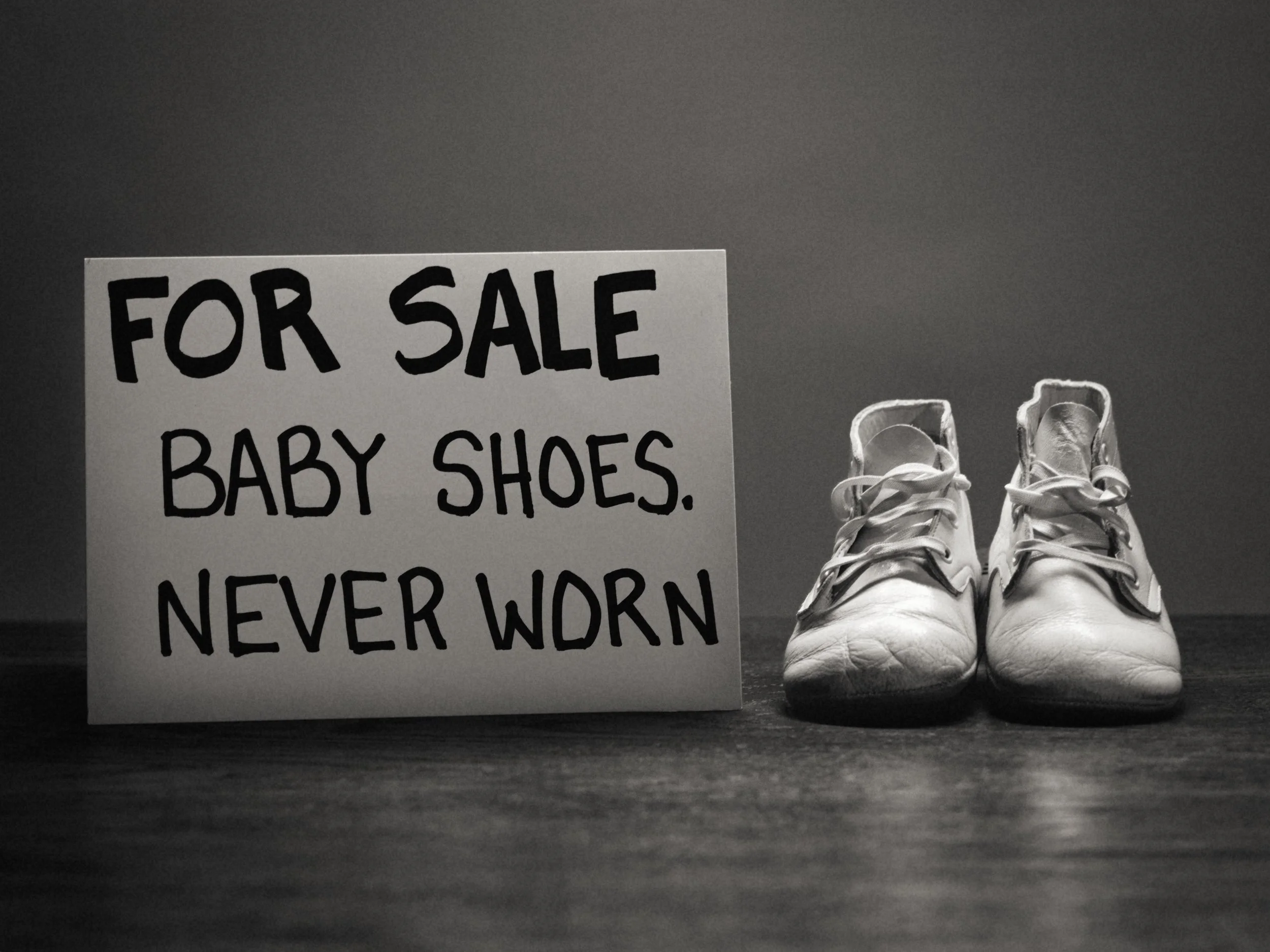A pair of white sneakers and a handwritten sign that reads ’For Sale Baby Shoes Never Worn’ against a dark background.