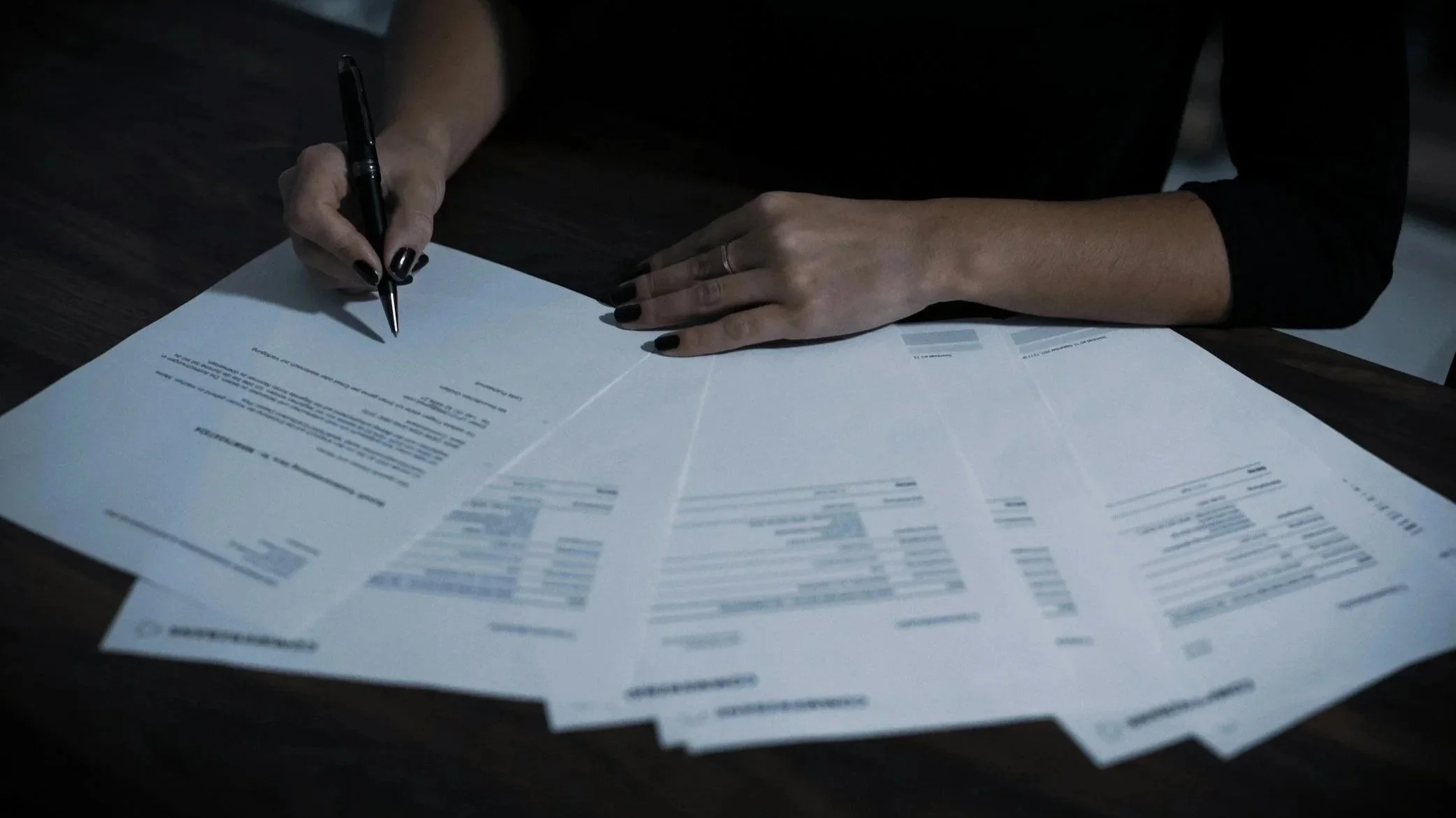 Person with dark nail polish signing documents on a dark wooden desk.