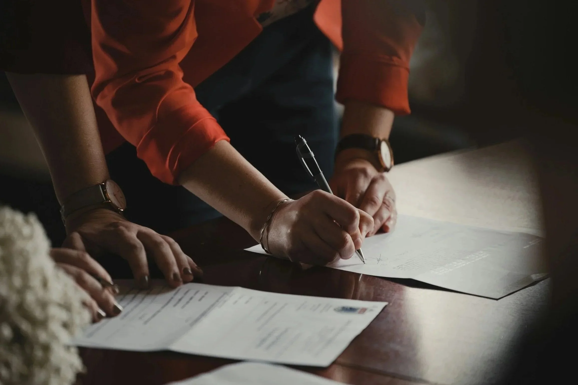 Multiple people are gathered around a table, writing on a sheet of paper. The focus is on their hands and arms, with watches and bracelets visible. The scene appears to be in a dimly lit room.