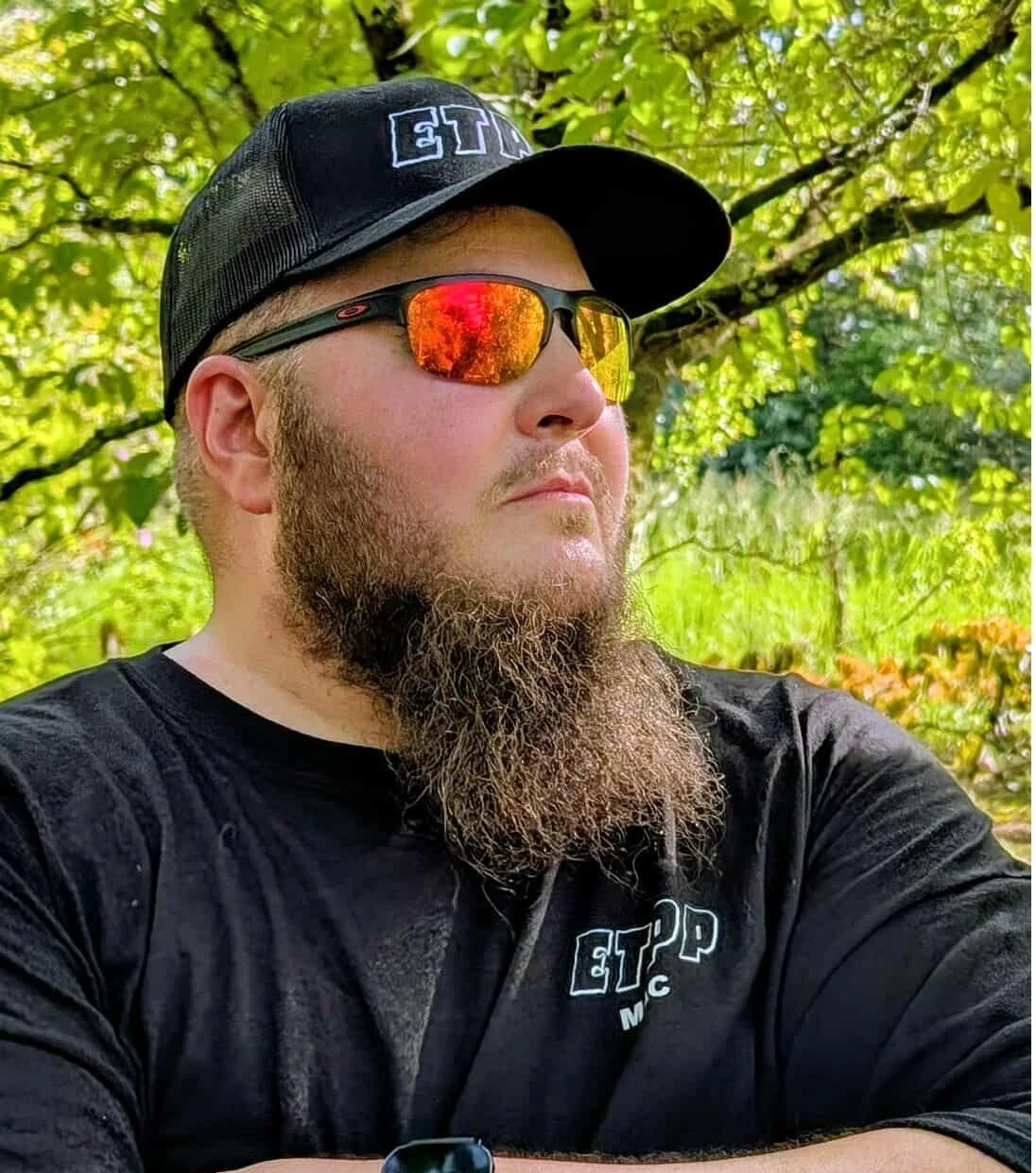 A man wearing a black cap and orange mirrored sunglasses, with a thick beard, outdoors with green trees in the background.