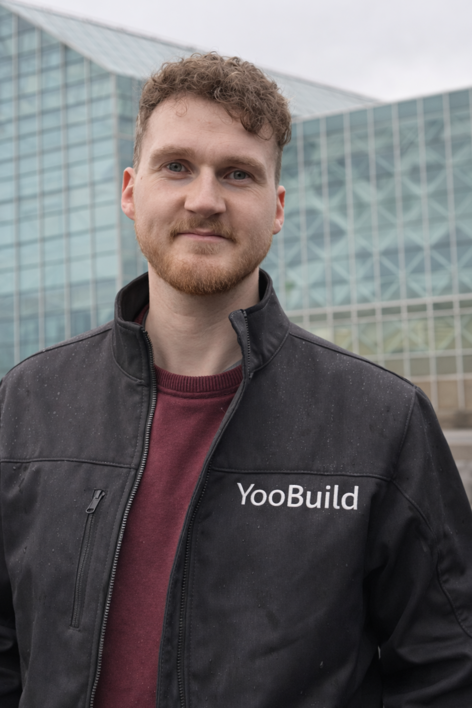 A young man with curly brown hair and a beard, wearing a black YooBuild jacket and a maroon shirt, standing outdoors in front of a modern glass building.