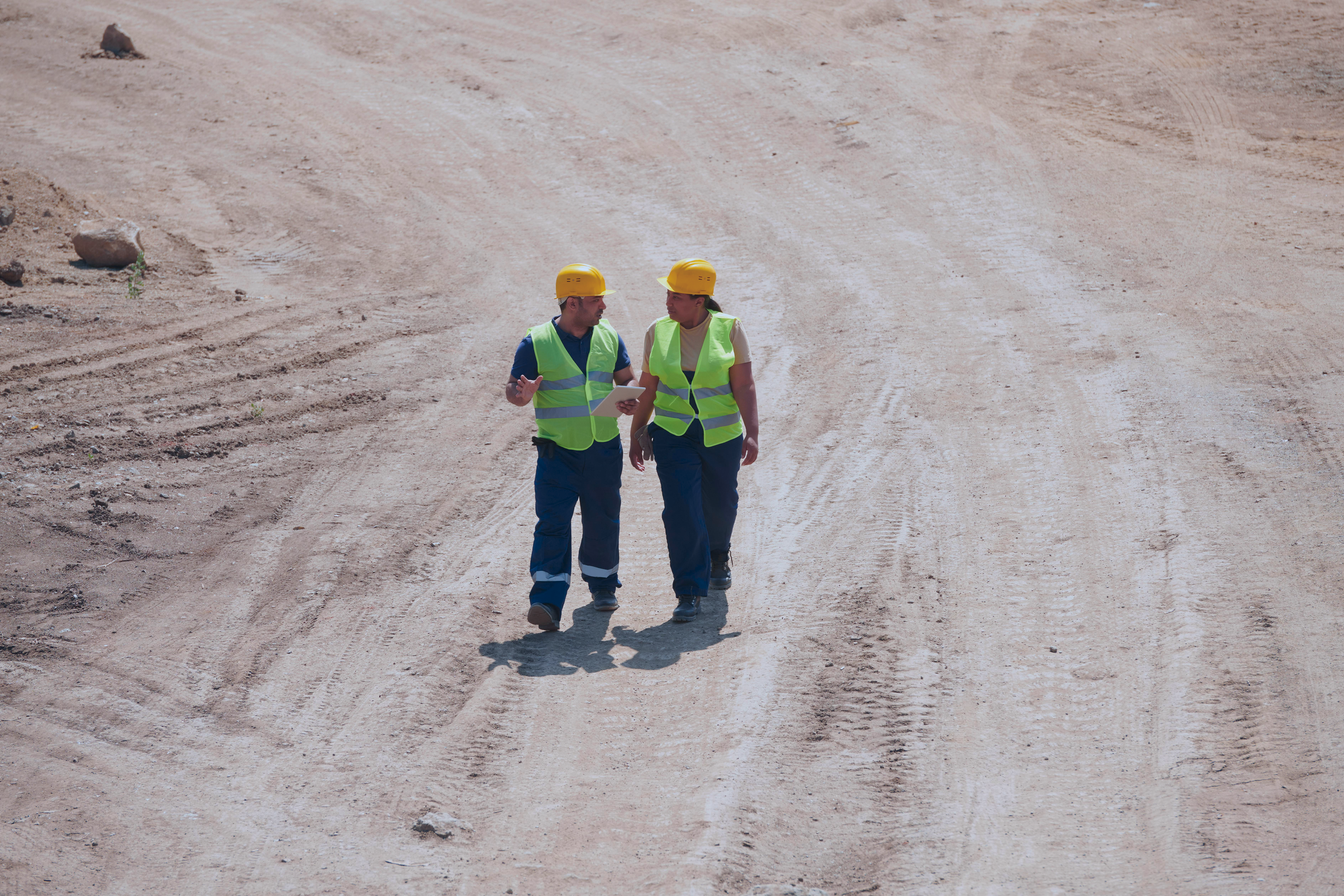 Two construction workers wearing yellow safety helmets and high-visibility vests walking and talking on a dirt road at a construction site.
