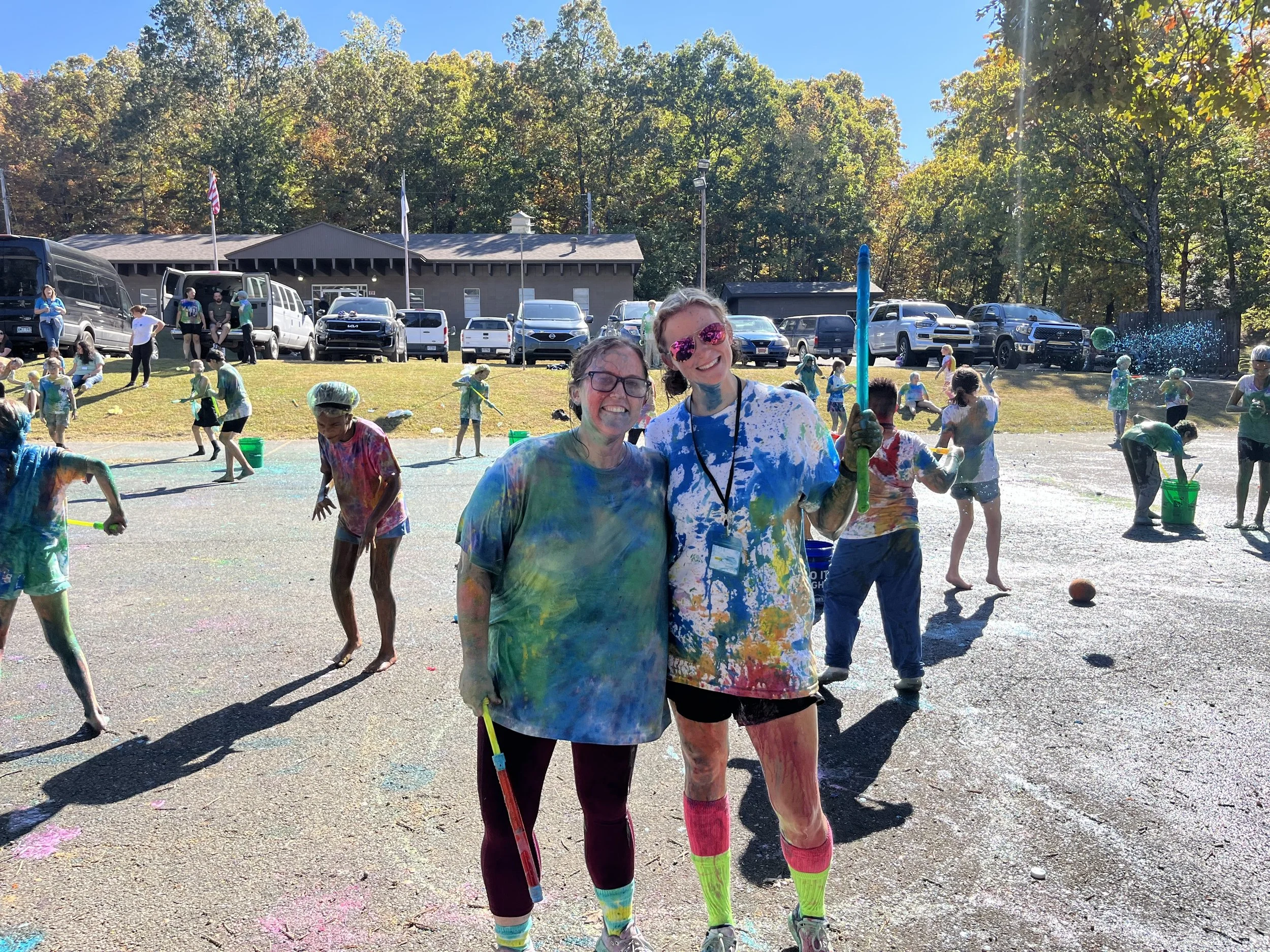 Two women smiling and covered in colorful powder during a color run event, with children playing and throwing powder in the background on a sunny day.