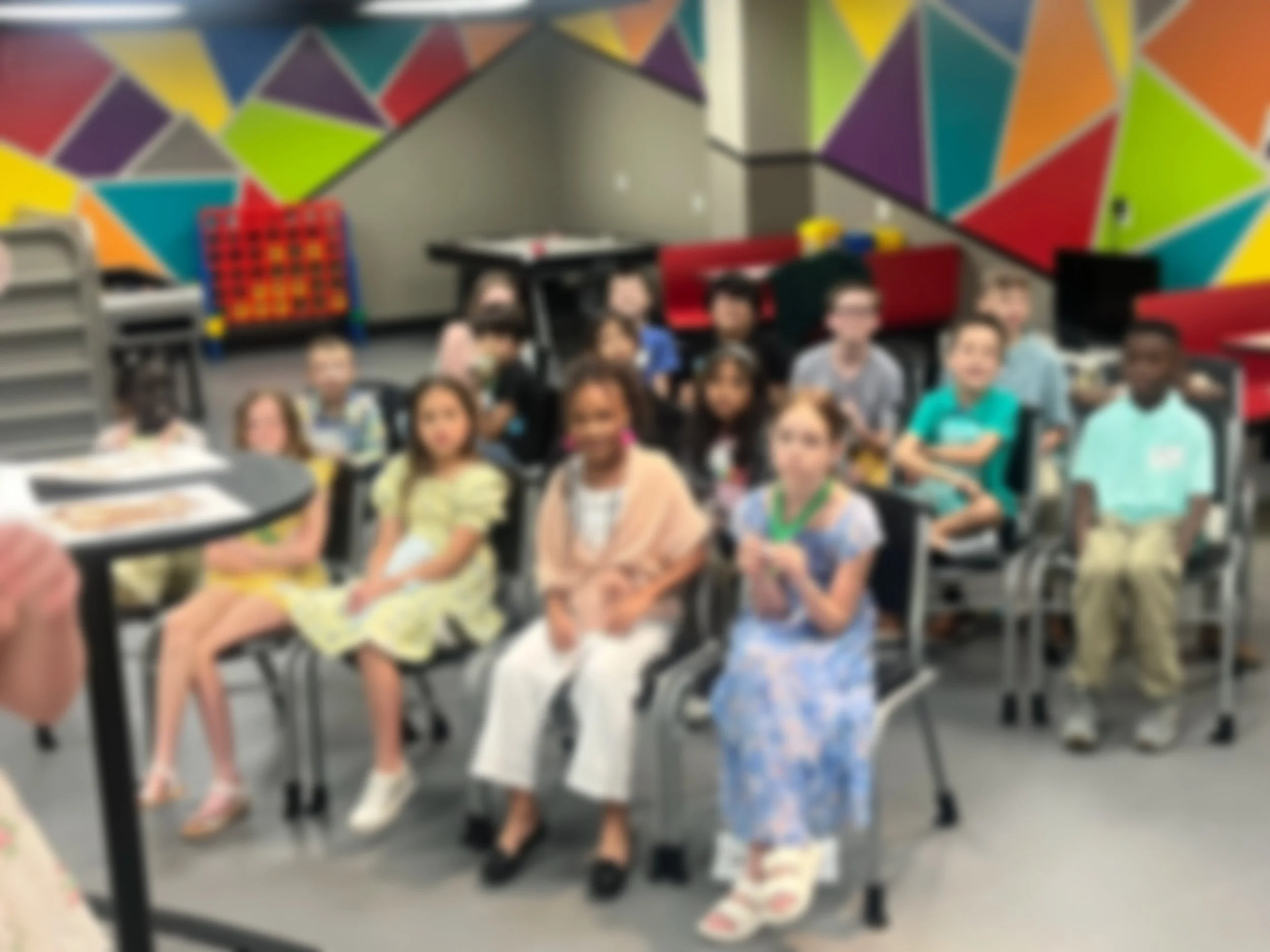 A group of children sitting in a classroom with colorful geometric wall art.