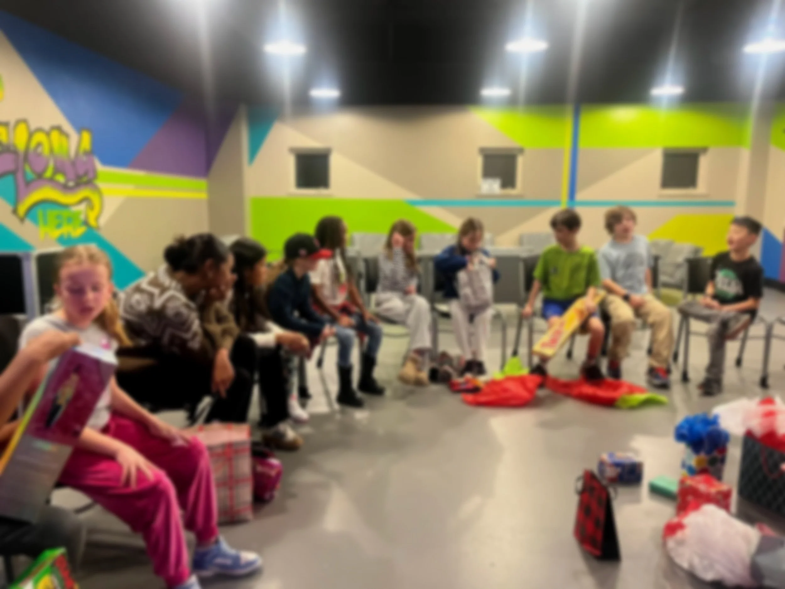 Children sitting in chairs in a colorful room with presents and other items on the floor, likely celebrating a party or gift exchange.