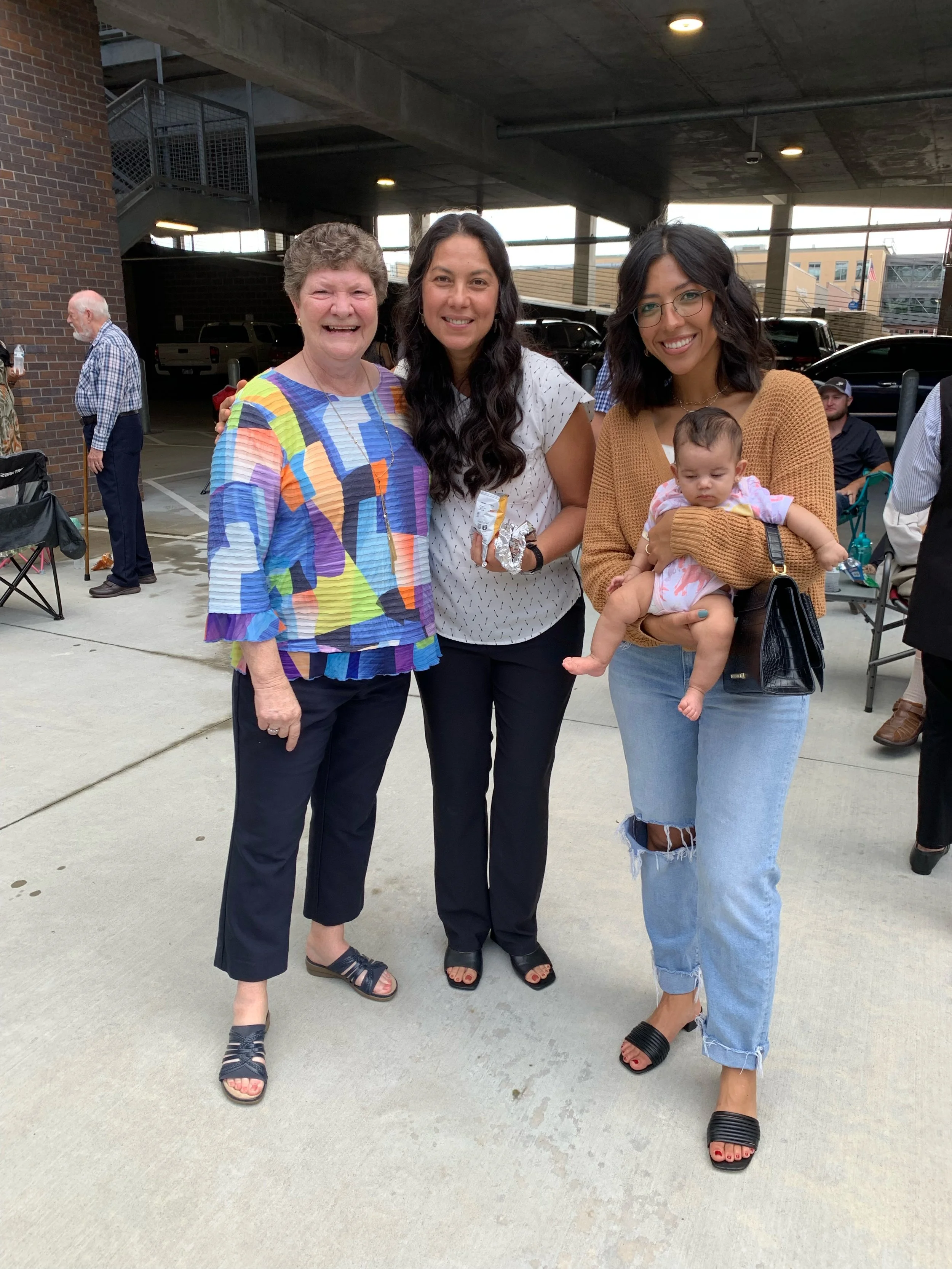 Four women standing together outside under a parking garage; one woman holding a baby girl; background includes cars and other people.