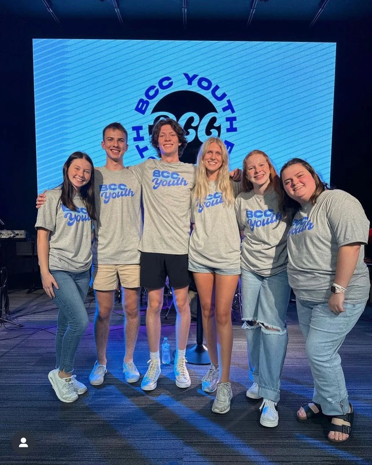 Group of six young people on stage, wearing matching gray T-shirts with blue text that says 'BCC Youth.' They are smiling and standing arm-in-arm. Behind them is a large screen with the logo 'THBCC YOUTH' in a circular design.
